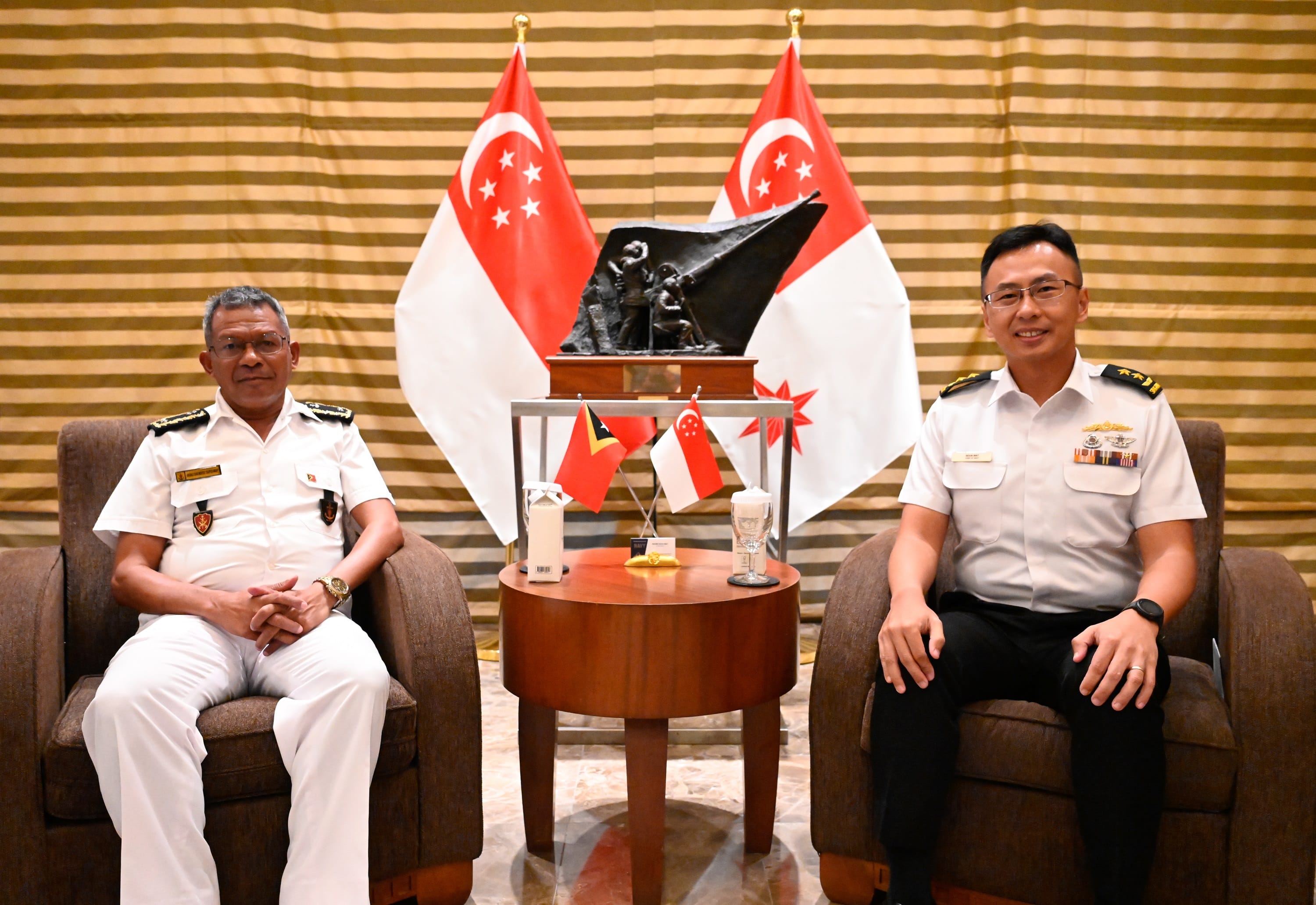 Two uniformed officers seated with Singapore flags and a sculpture in the background.