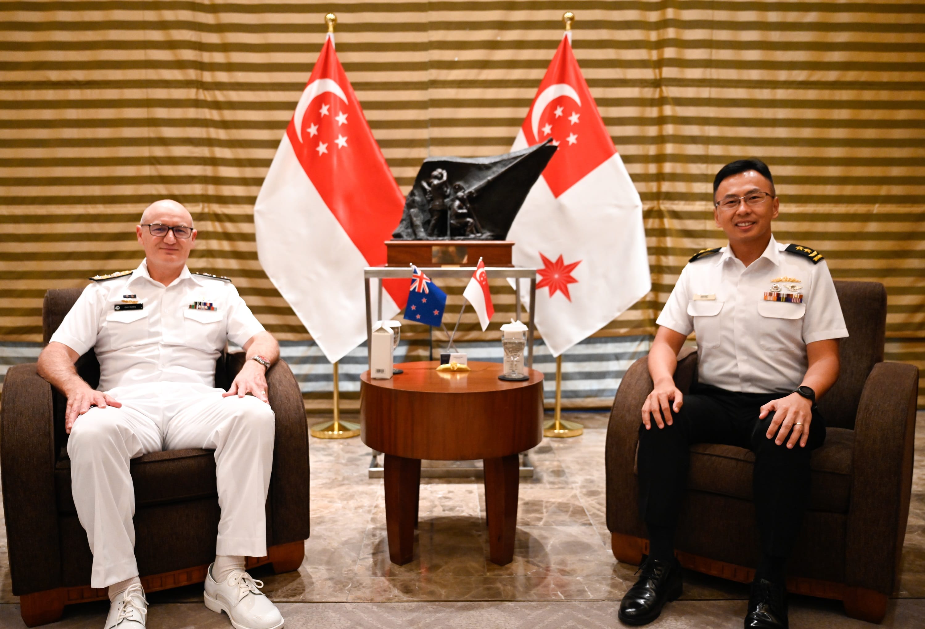 Two men in naval uniforms seated, Singaporean flags behind. Table has New Zealand and Singapore flags.