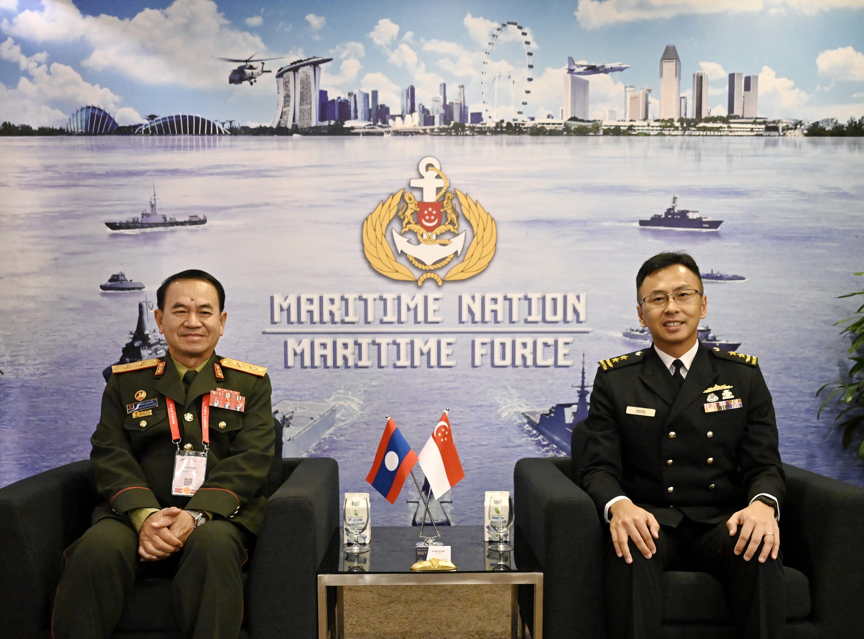 Two naval officers seated, with flags of Lao PDR and Singapore in front, backdrop of Maritime Nation Maritime Force.