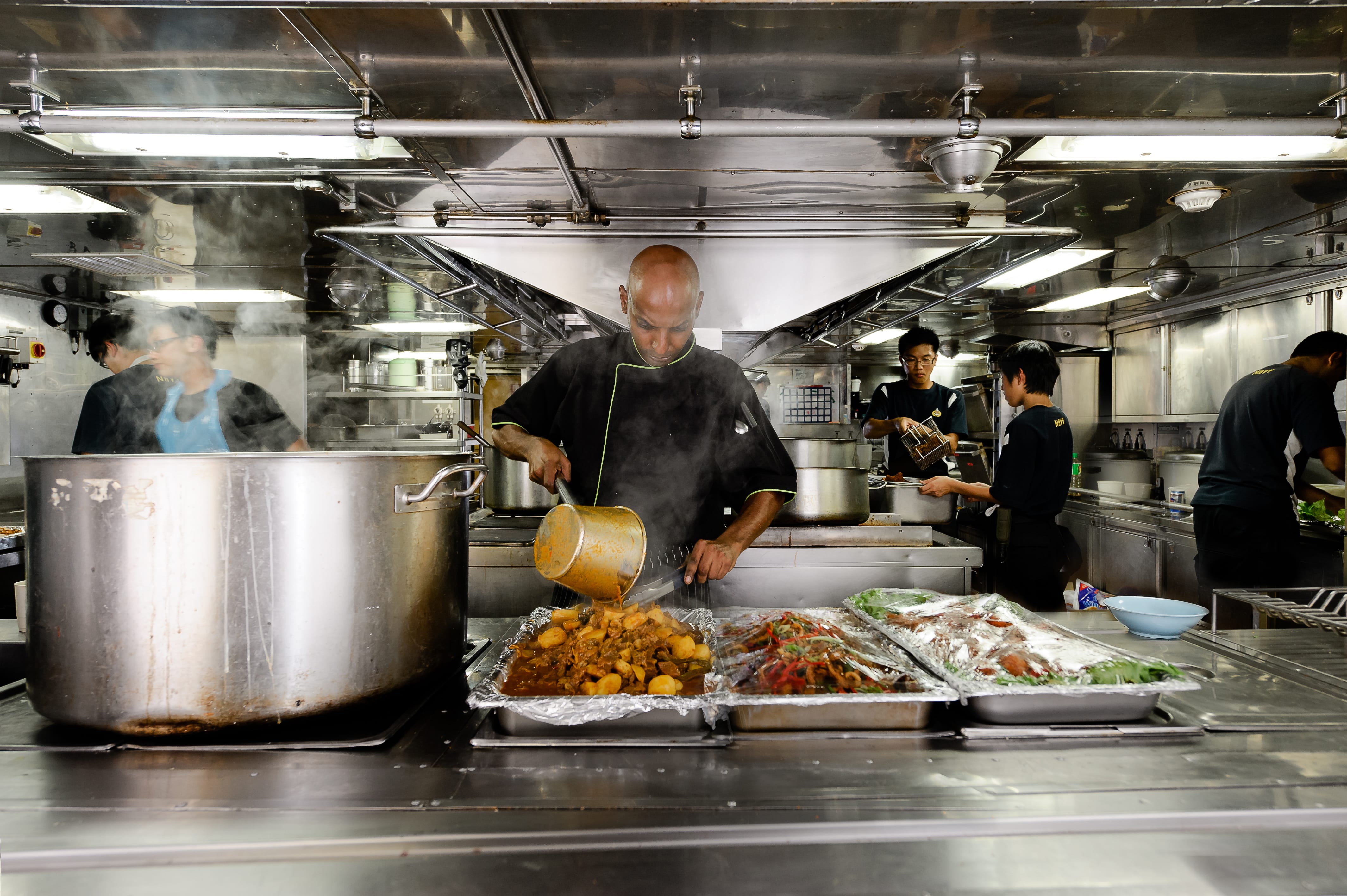 Crew members prepare and cook meals in a large, steaming kitchen aboard a ship.