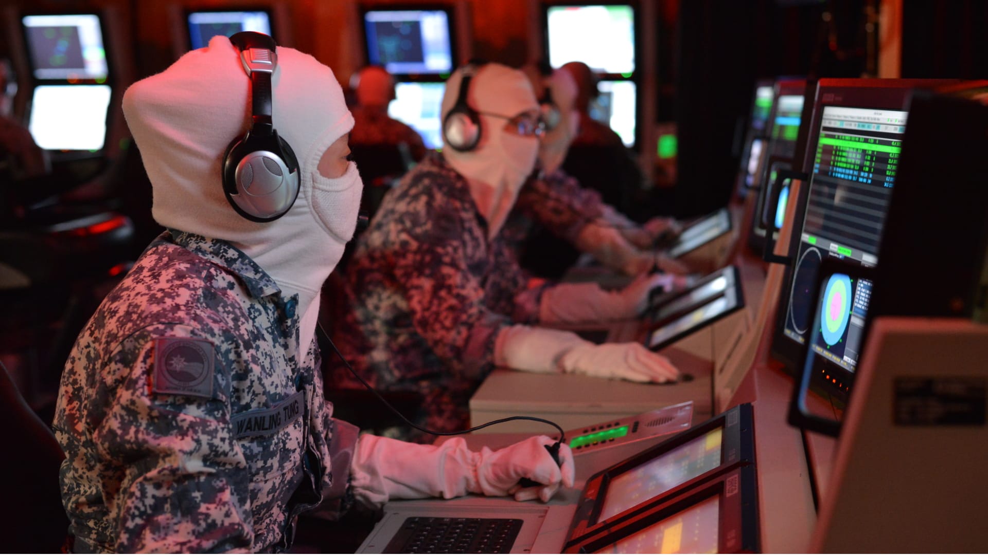 Naval crew in protective gear operate radar and control systems in a dark command center.