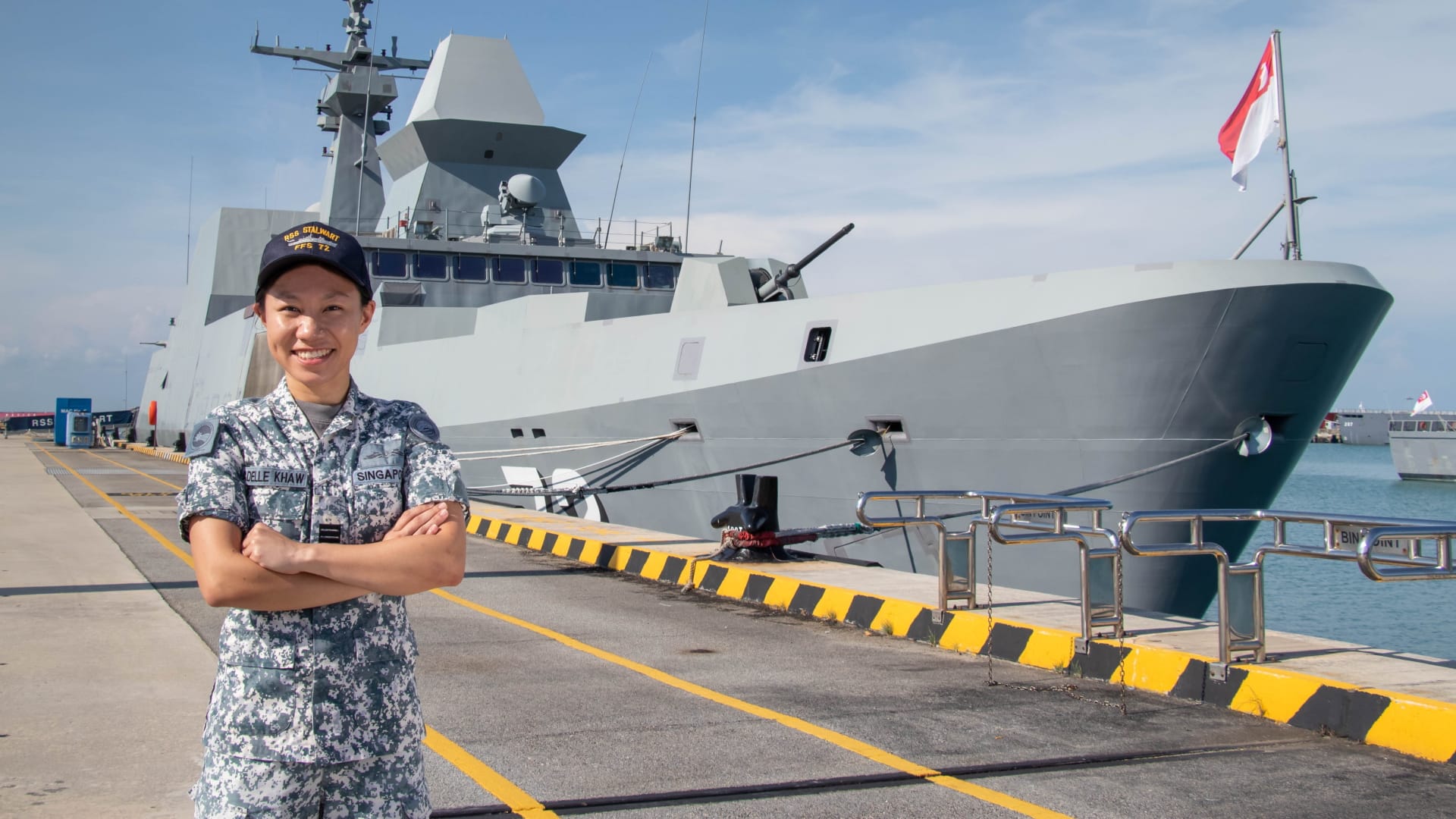 CPT Adelle Khaw stands smiling in uniform in front of RSS Stalwart at a naval pier.