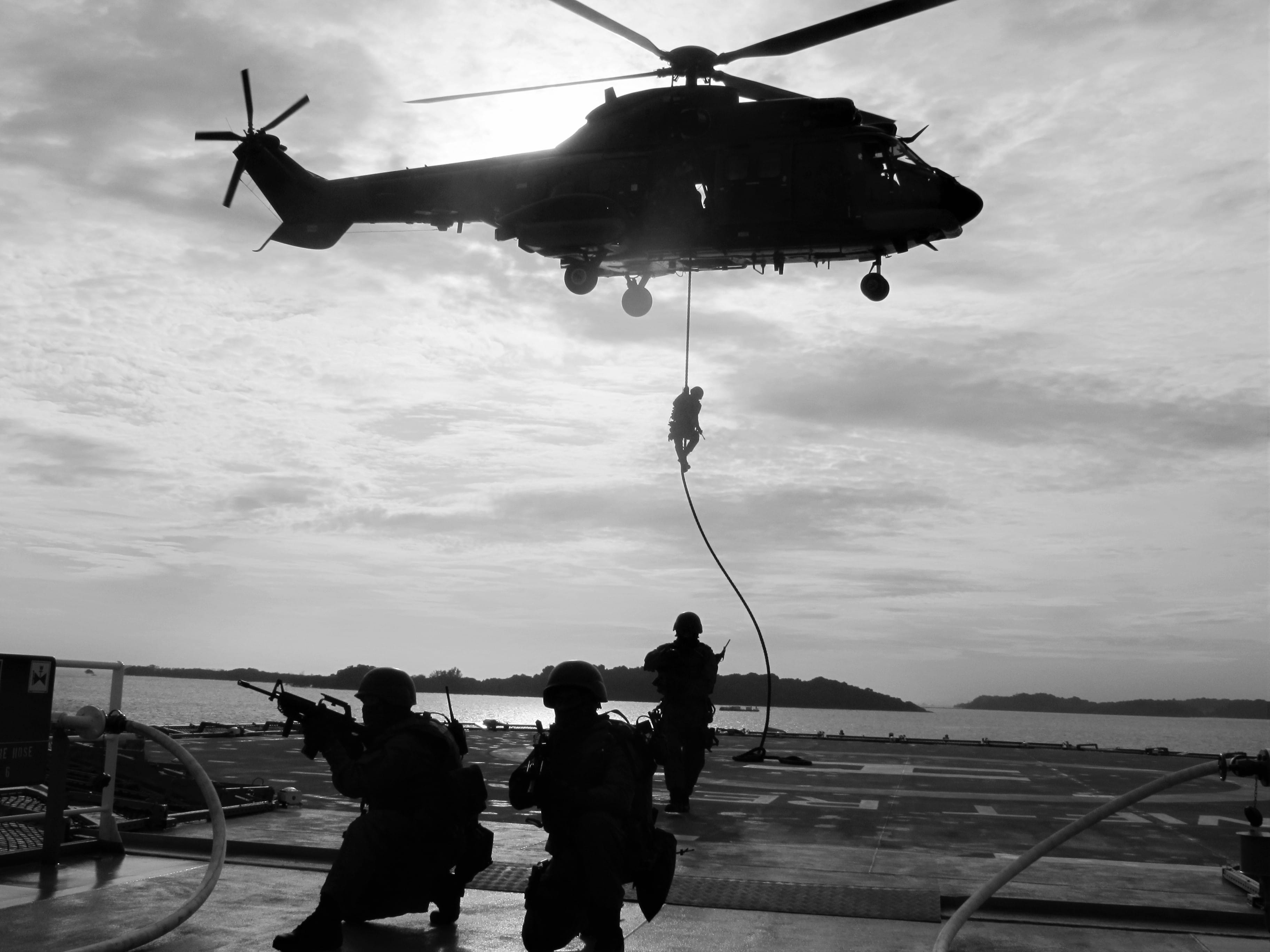 A navy helicopter performing a rope descent exercise on a naval ship deck.