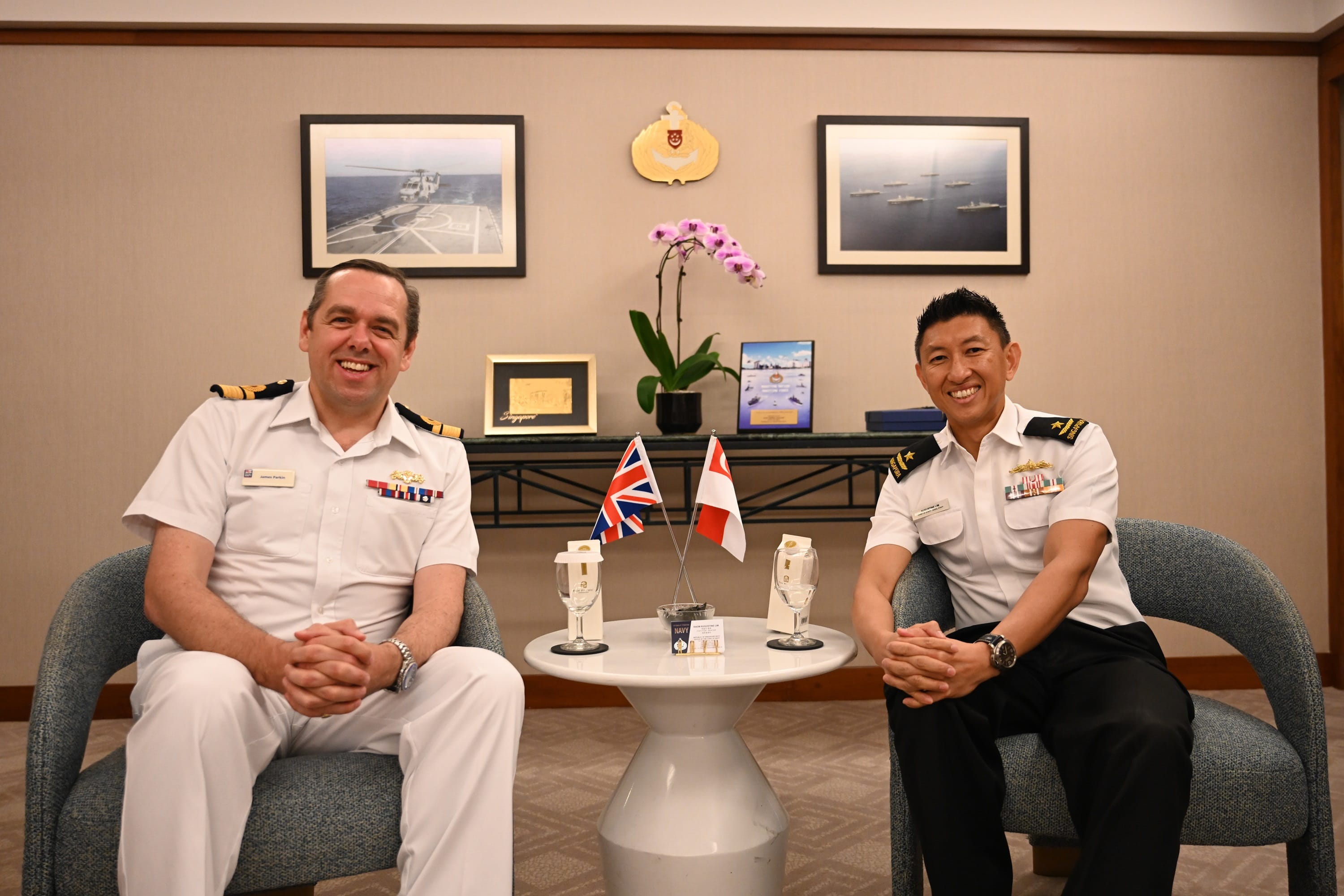 Two naval officers in uniform seated, smiling near table with UK and Singapore flags.