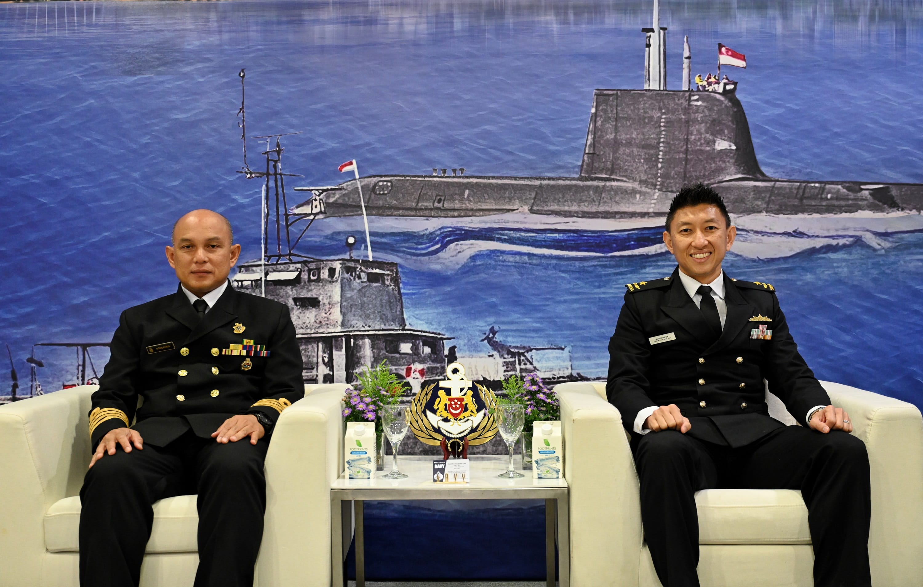 Two naval officers from Brunei and Singapore seated in front of an illustrated maritime backdrop.