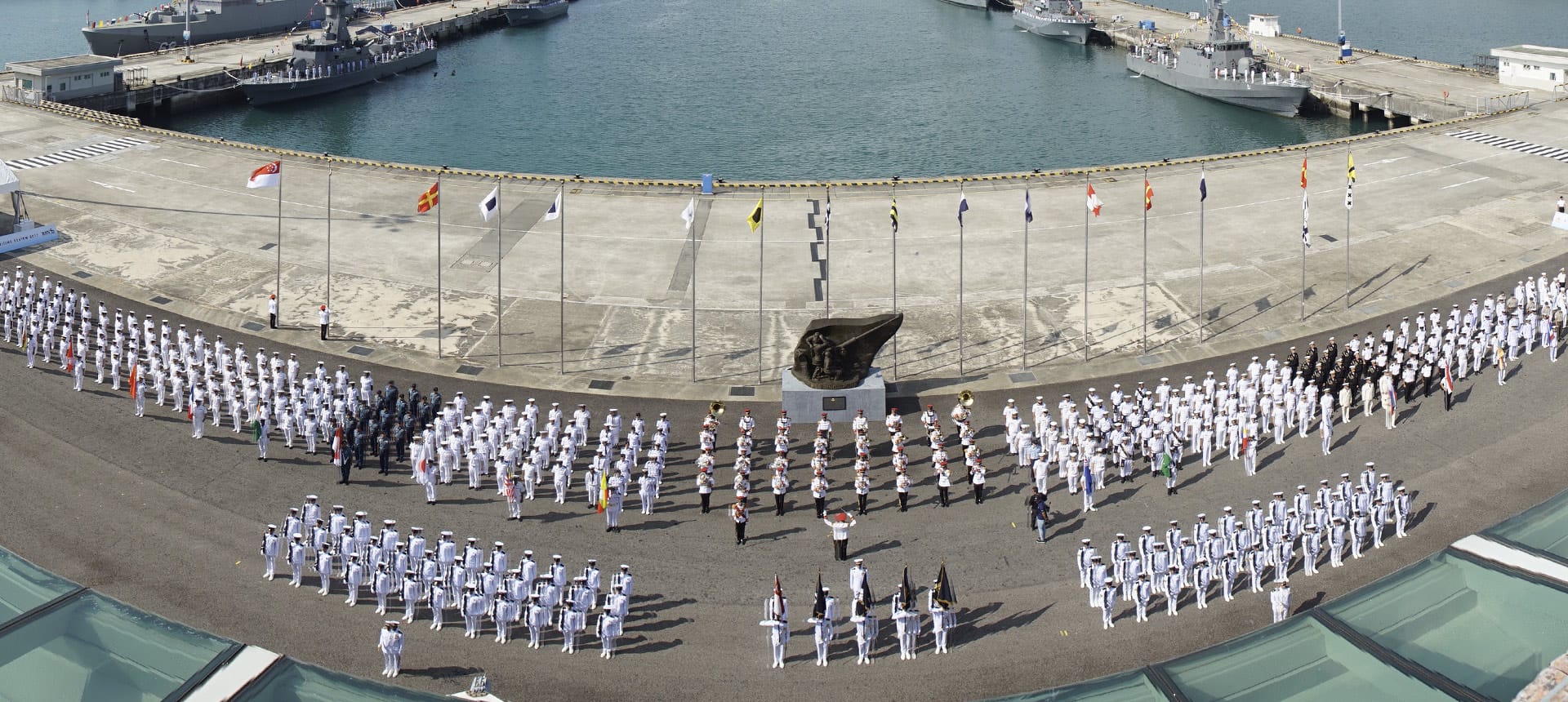 Naval personnel from various countries stand in formation at a dockside parade with flags representing participating nations.
