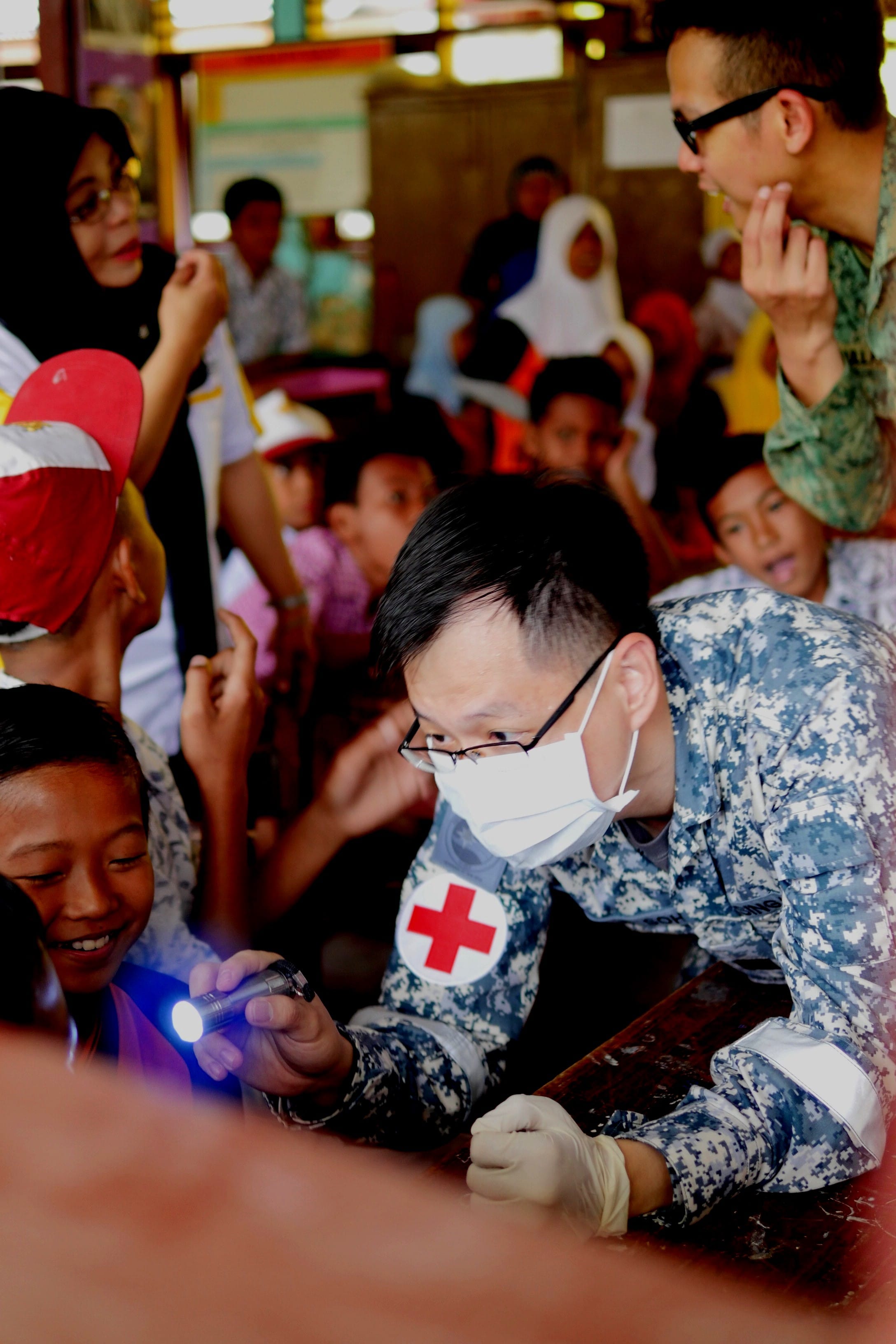 A navy medic in a mask and gloves examines a child with a flashlight during a humanitarian mission.