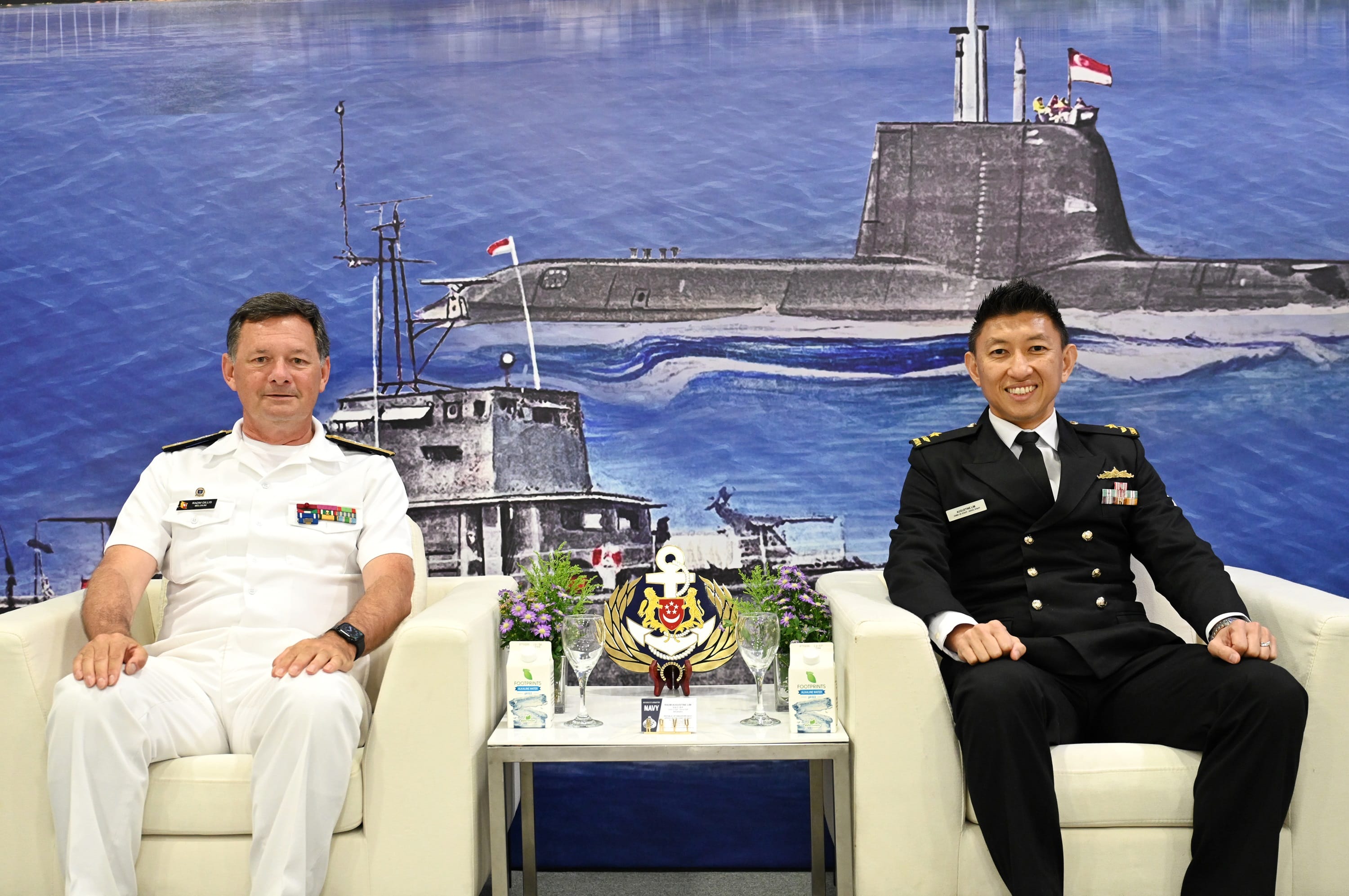 Two naval officers from Belgium and Singapore seated in front of an illustrated maritime backdrop.