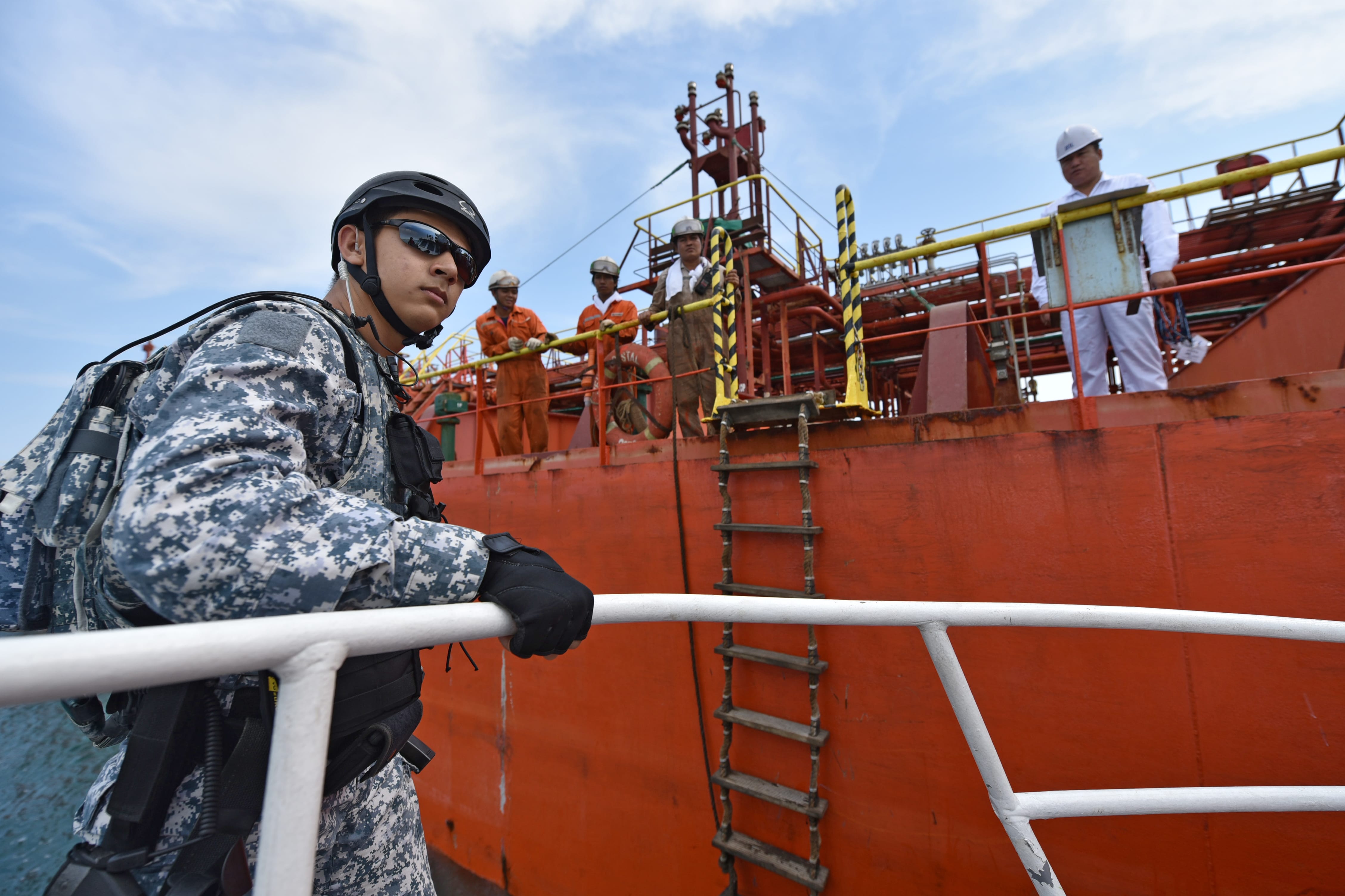 A navy personnel in helmet and sunglasses approaches a commercial vessel where crew members await at the deck.