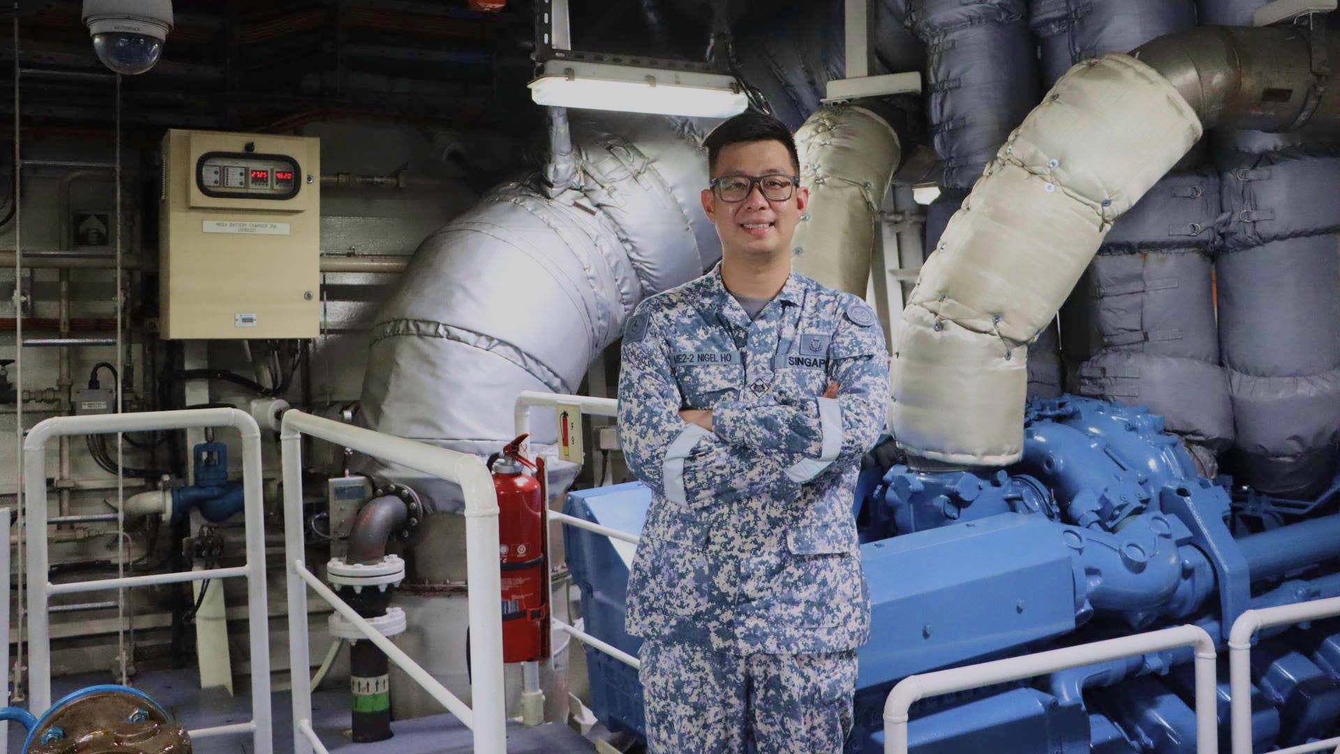 ME2 Nigel Ho stands confidently in a ship’s engine room, surrounded by large insulated pipes and machinery.e