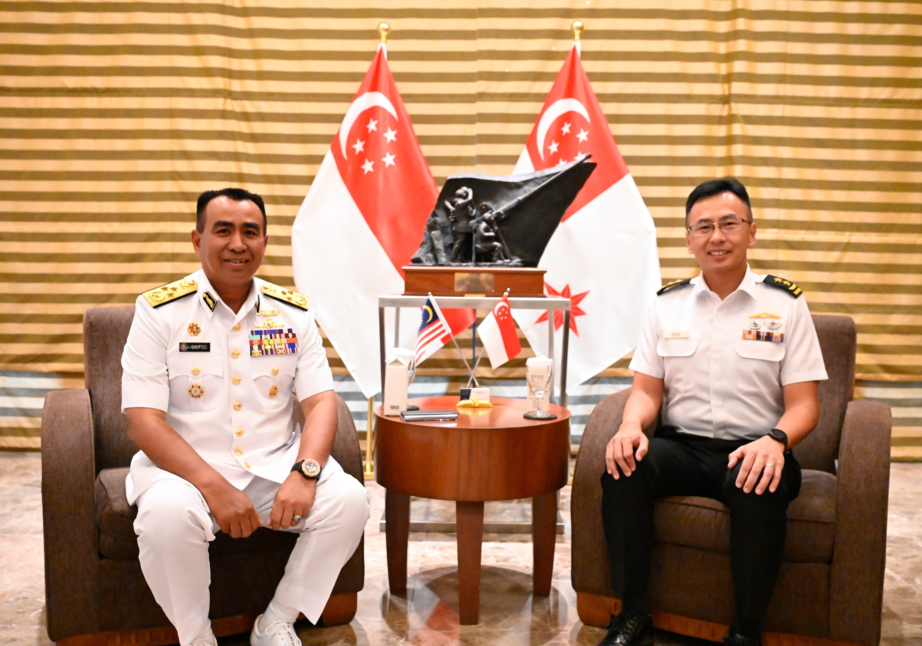 Two people in naval uniforms pose in armchairs with Singapore flags behind them.