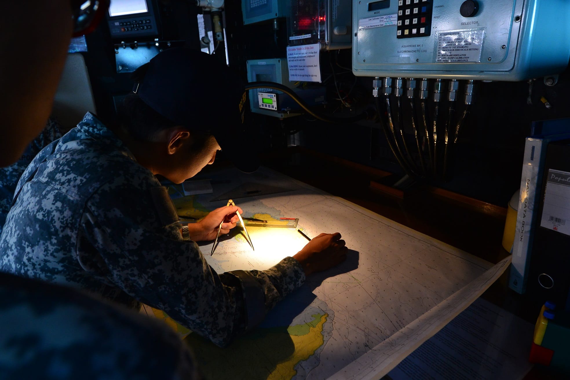 A navy personnel at a desk, holding a compass in one hand, paper charting.