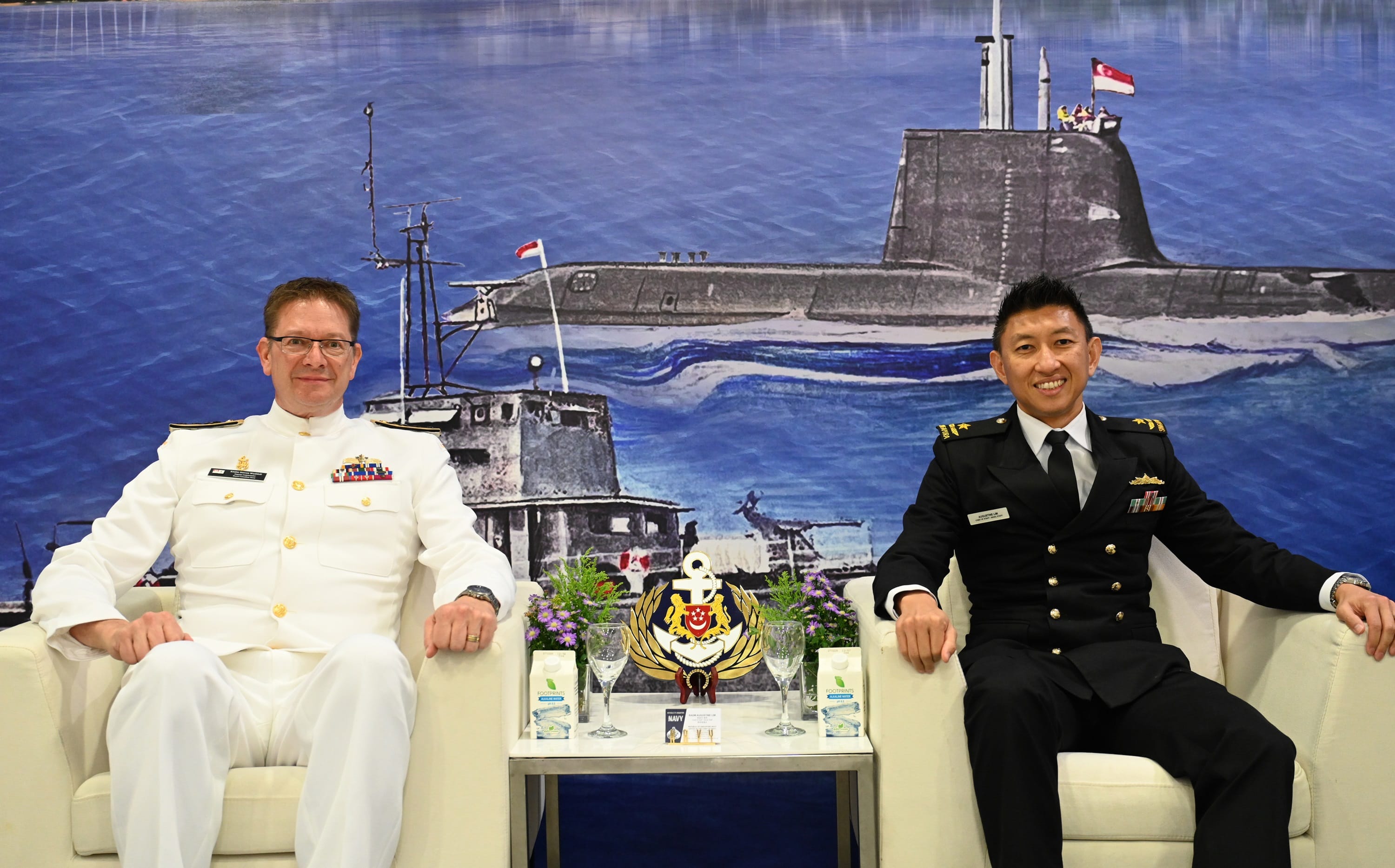 Two naval officers from Canada and Singapore seated in front of an illustrated maritime backdrop.