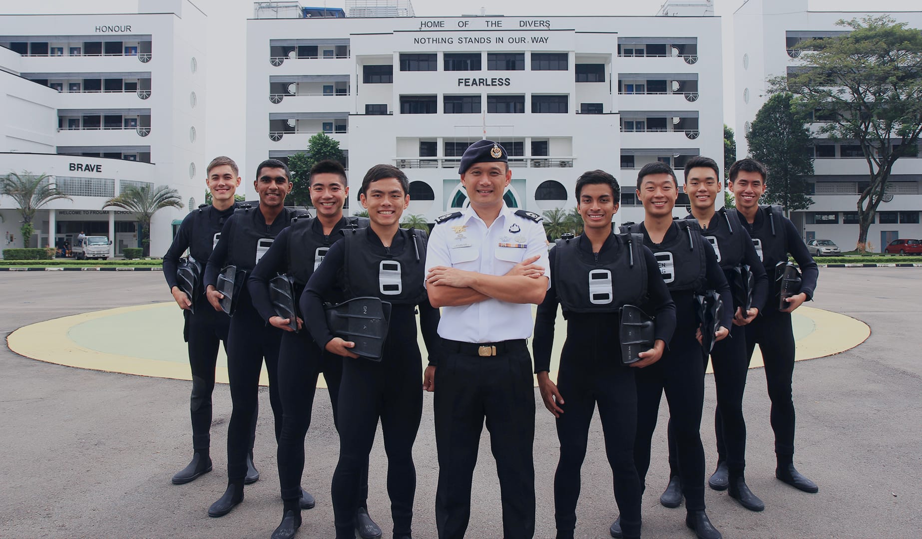 MWO in white naval uniform stands with Navy divers in wetsuits outside SAF diving school marked “FEARLESS.”