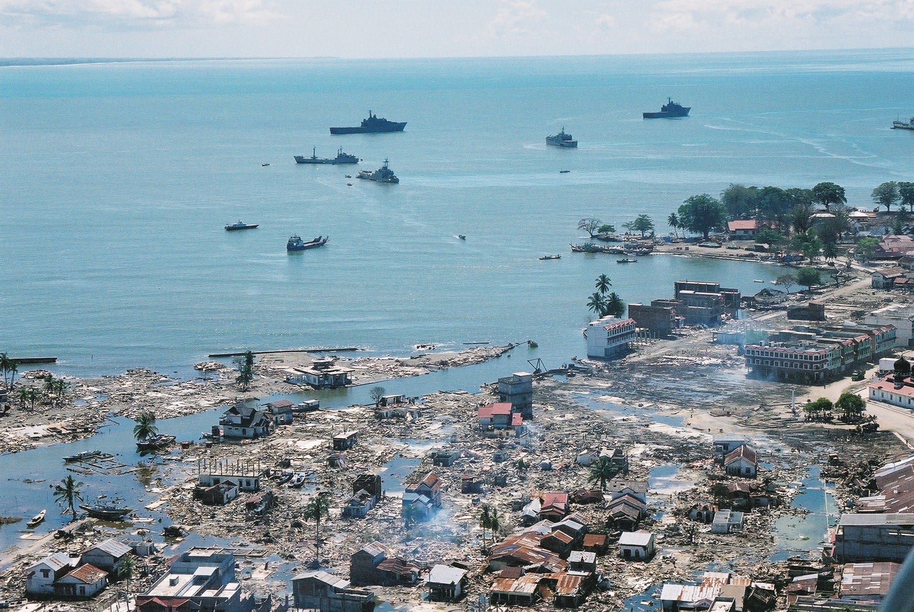 Aerial view of coastal city devastation; ships in ocean background.