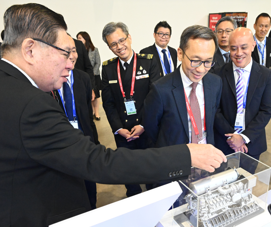 Men in suits and naval uniform examine a model engine in a clear display case.