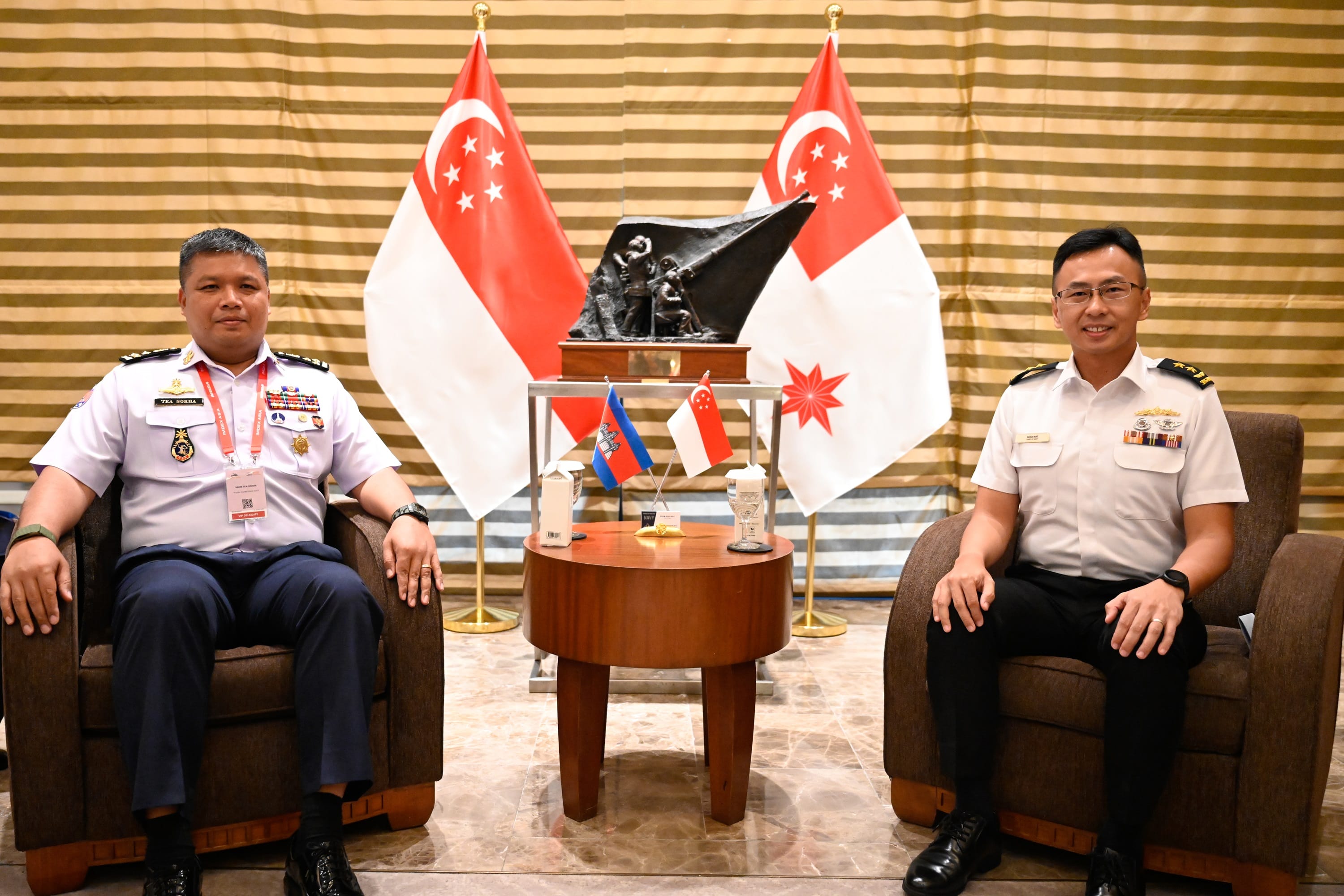 Two uniformed officers sit in chairs, flanked by Singapore flags. A brown table with mini flags is centered.