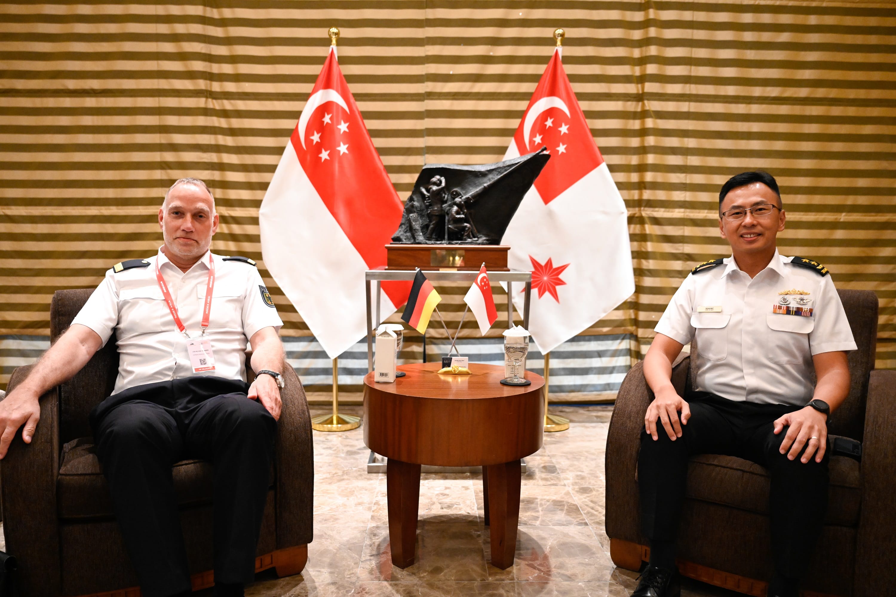Two men in uniform sit in chairs with Singapore flags behind them.