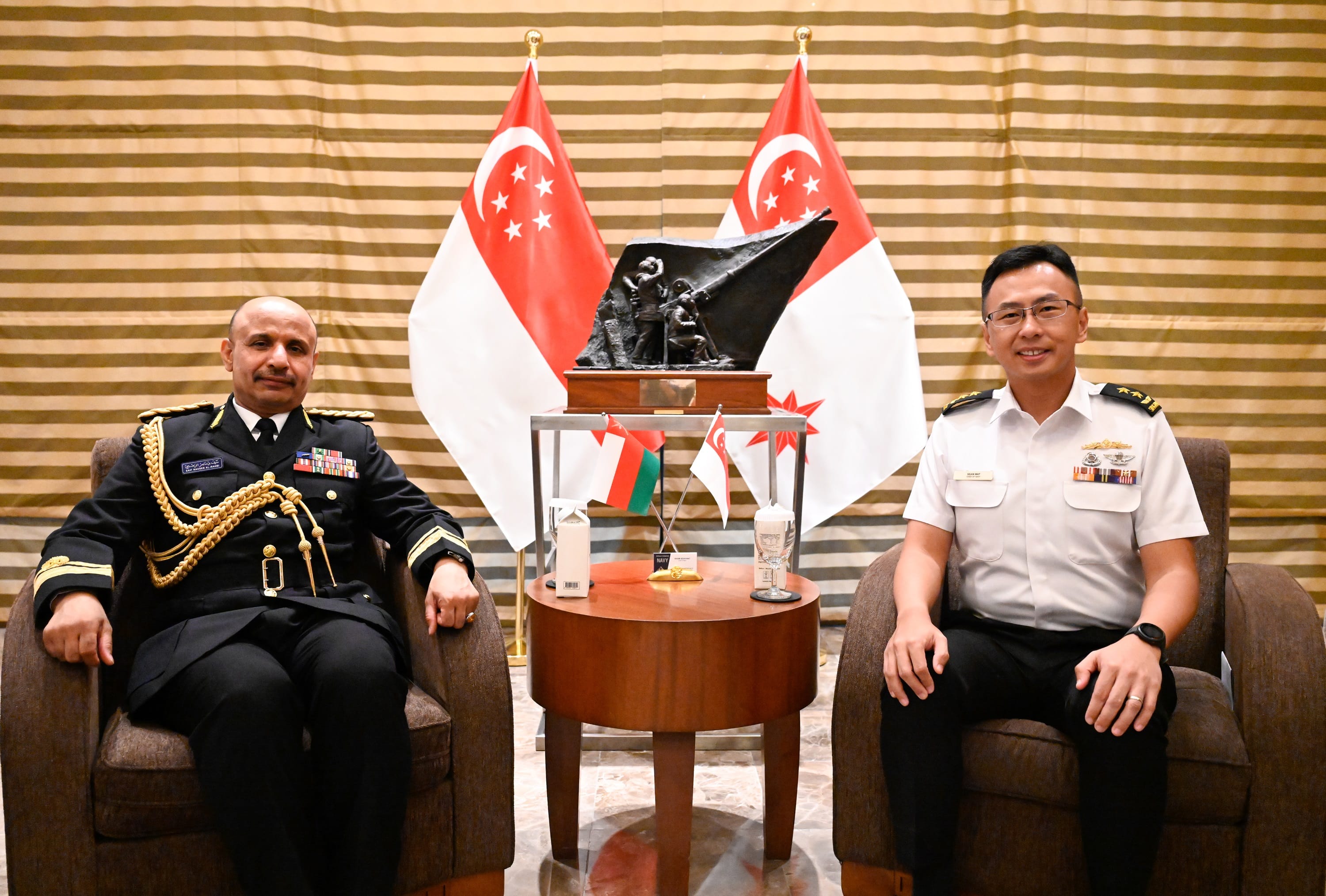 Two military officials seated, Singapore flags behind them with a striped wall.
