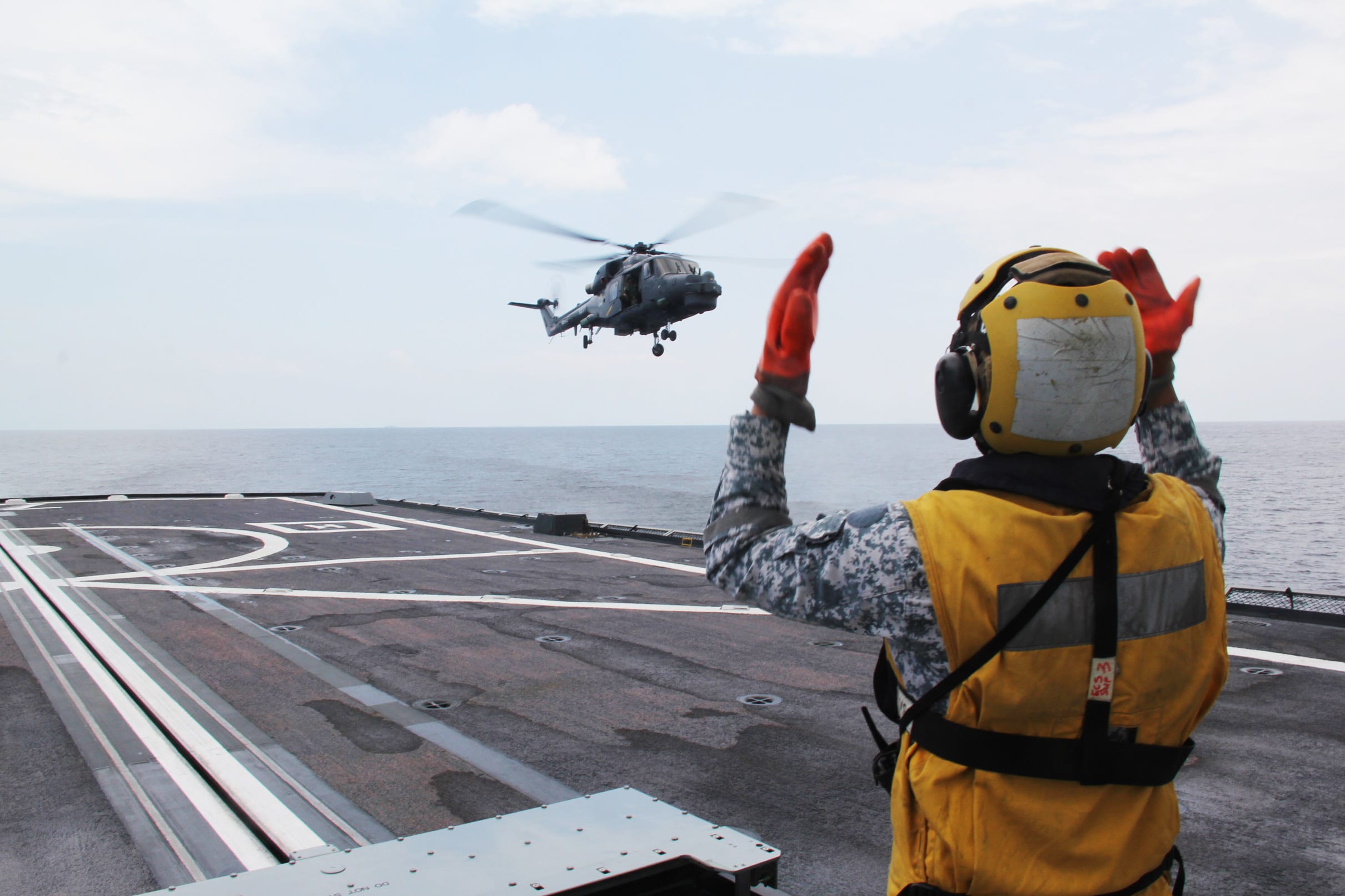 A naval crew member in a helmet and vest signals to a helicopter landing on a ship's flight deck.
