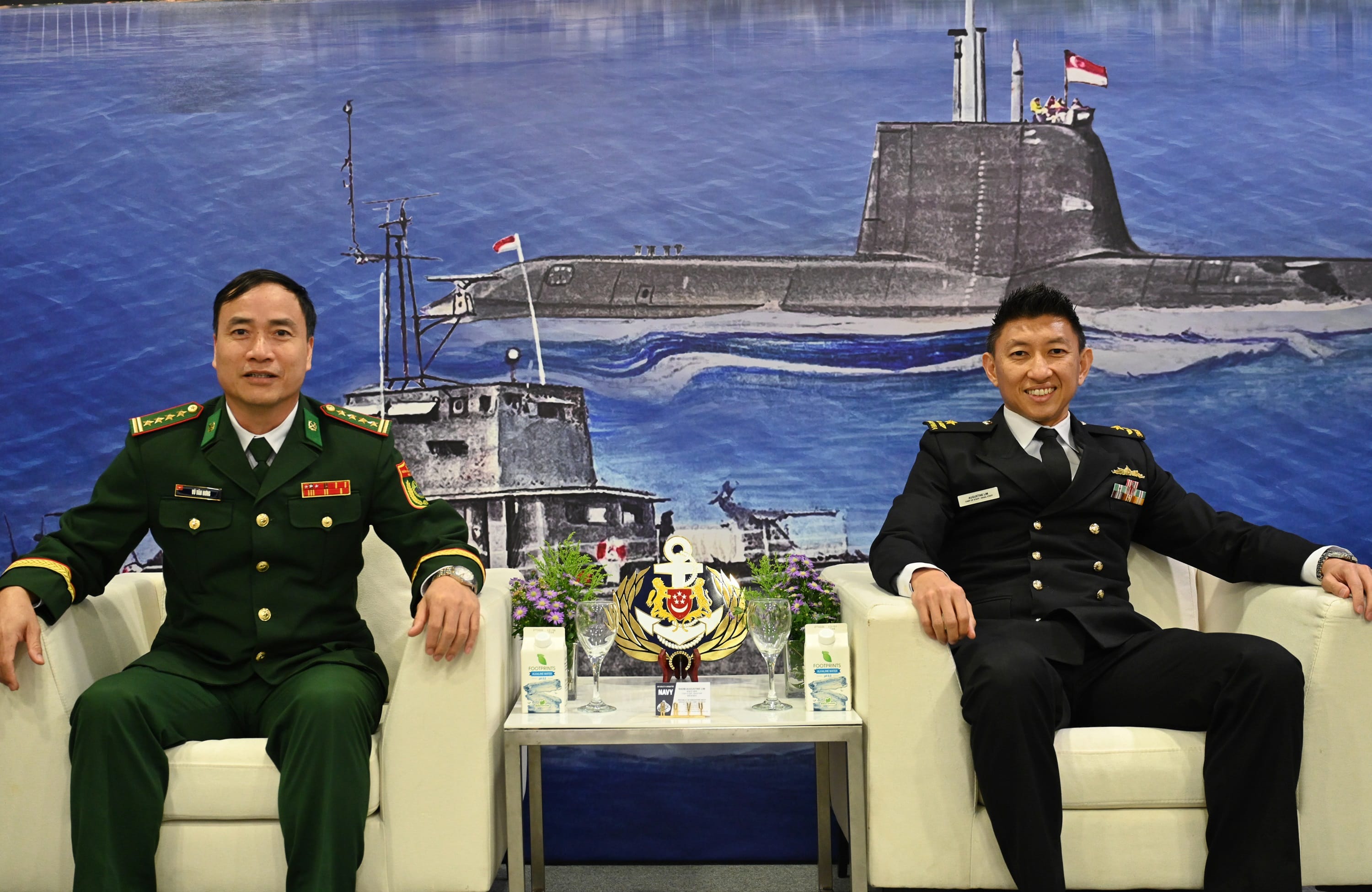 Two naval officers from Vietnam and Singapore seated in front of an illustrated maritime backdrop.