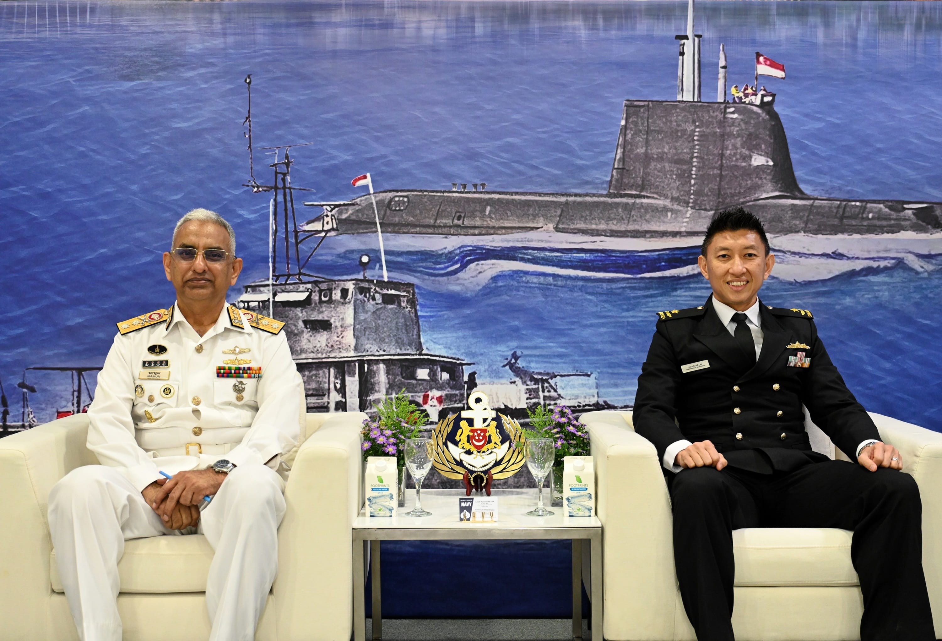 Two naval officers from Bangladesh and Singapore seated in front of an illustrated maritime backdrop.