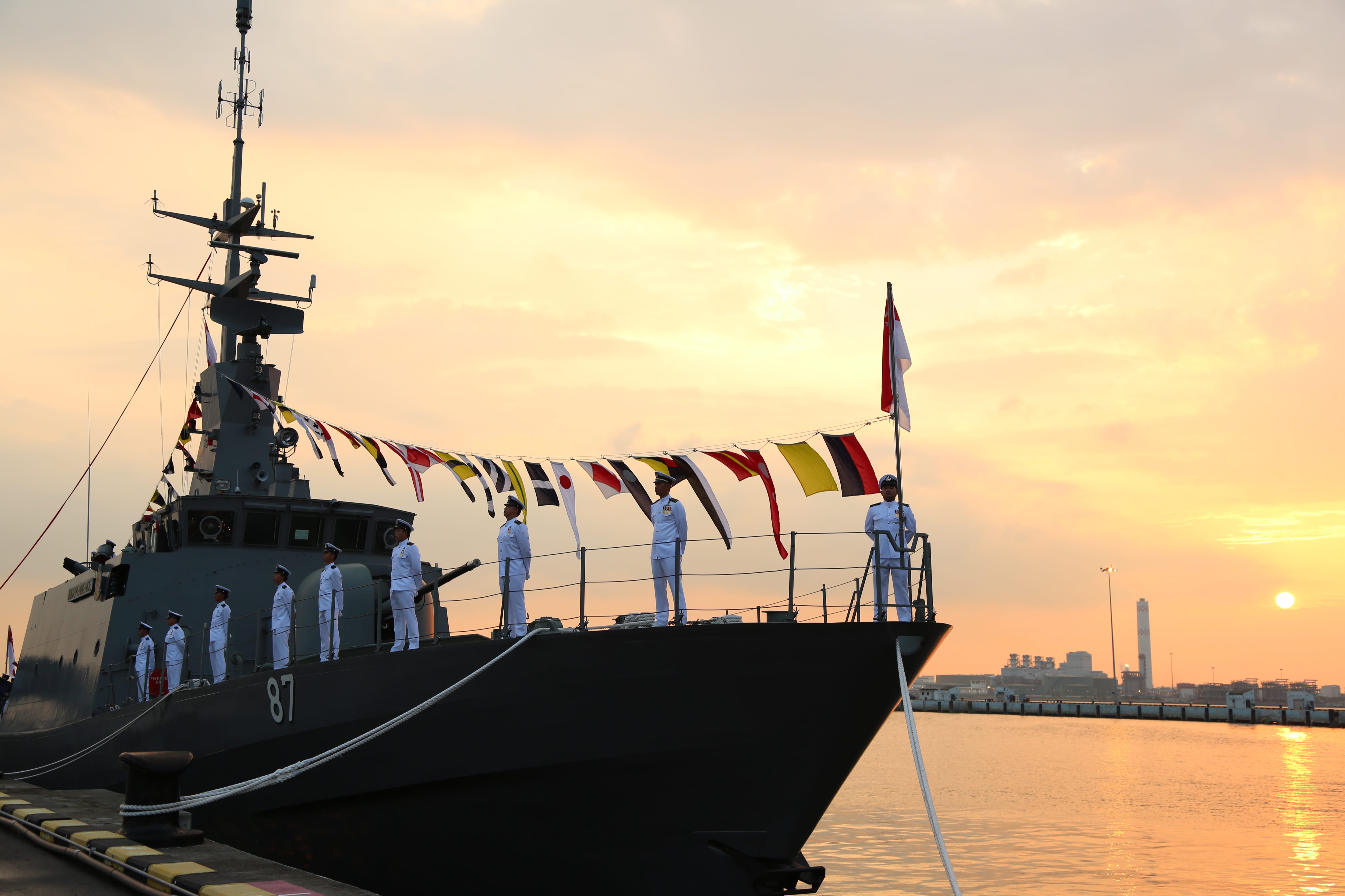 Navy personnel standing at attention on the bow of a vessel as it docks at sunset.