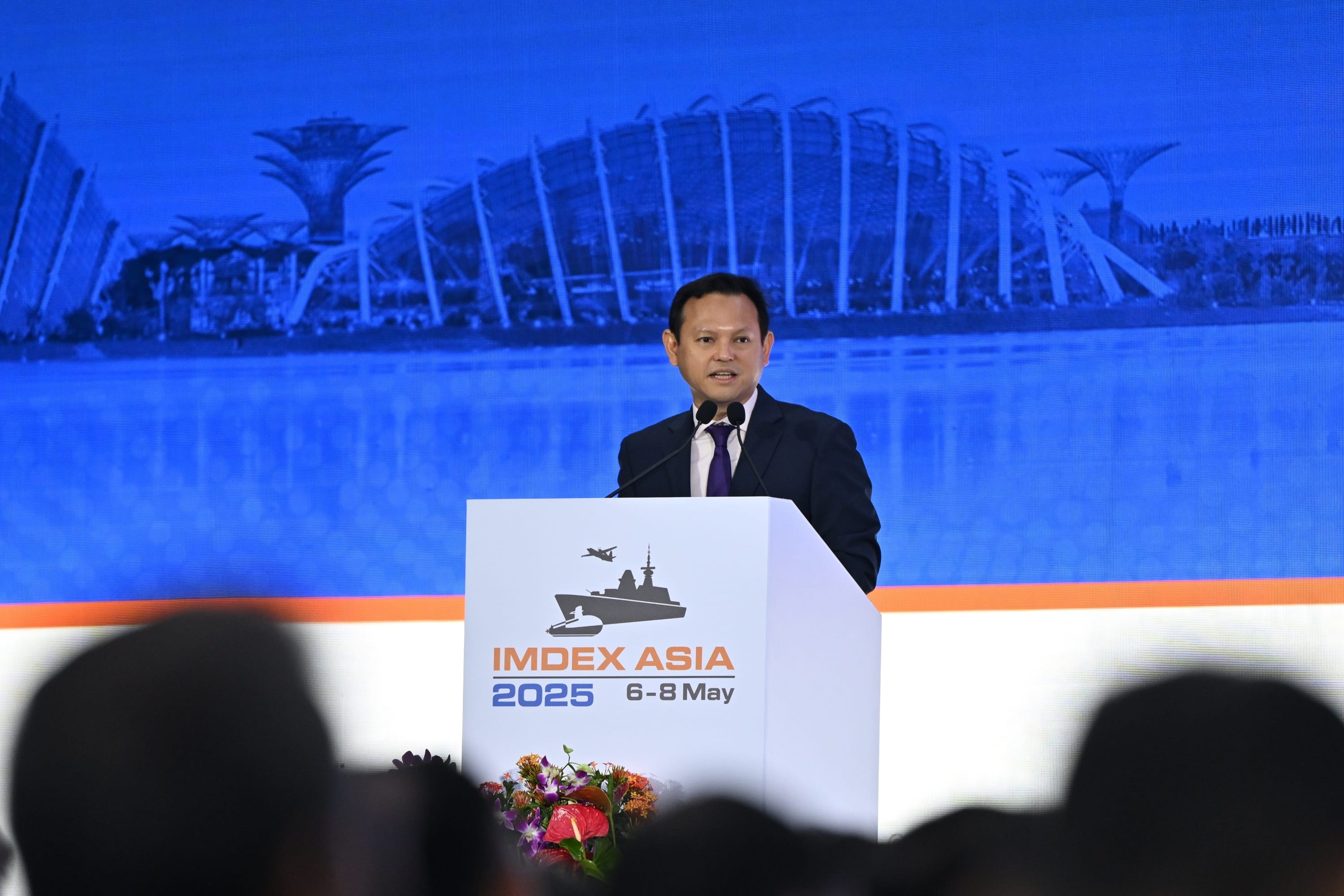 Speaker at podium with "IMDEX ASIA 2025" logo; gardens by the bay in background.