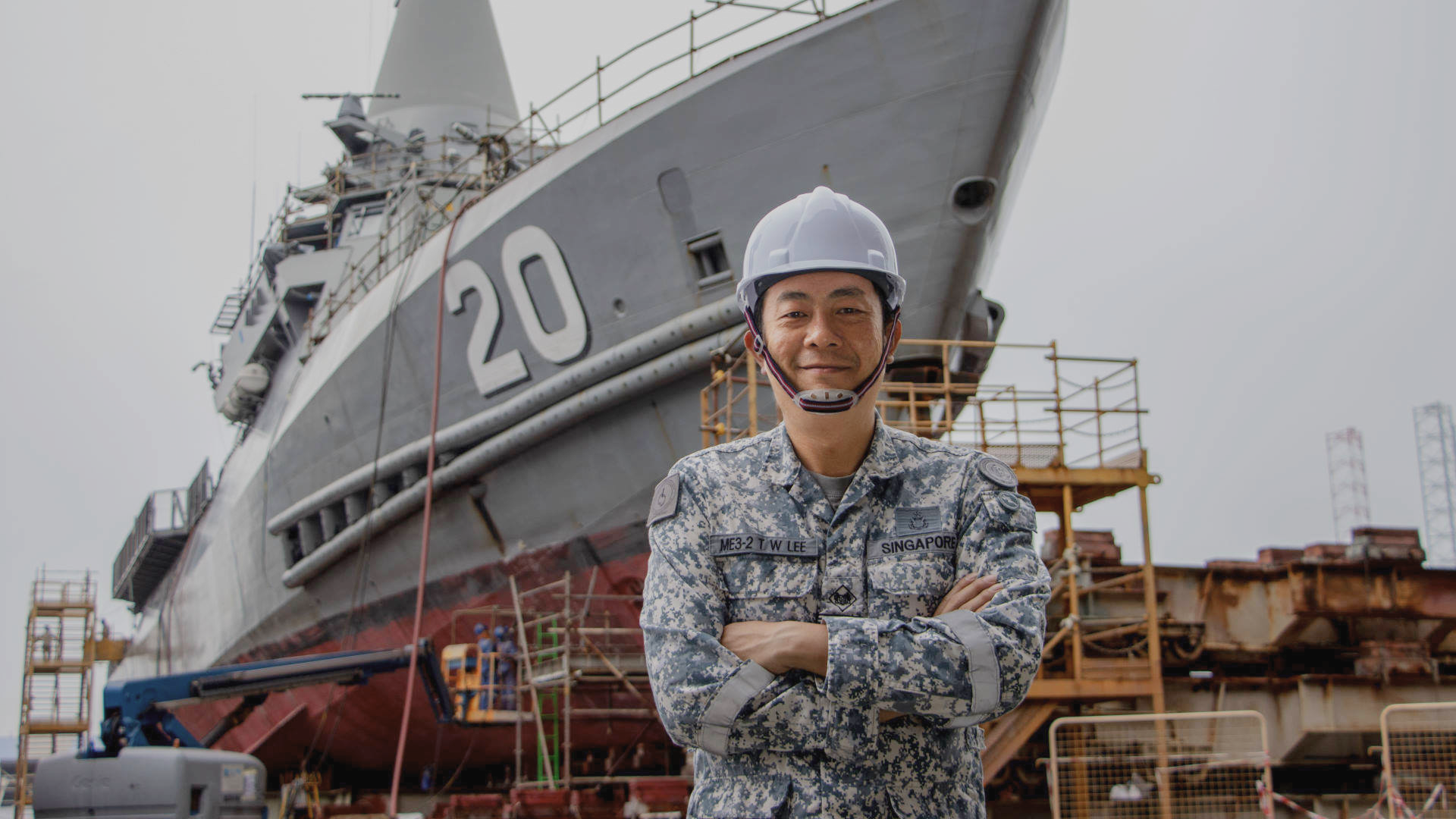 ME3 Lee Teck Wee in Navy uniform and helmet stands proudly in front of a docked warship under maintenance.