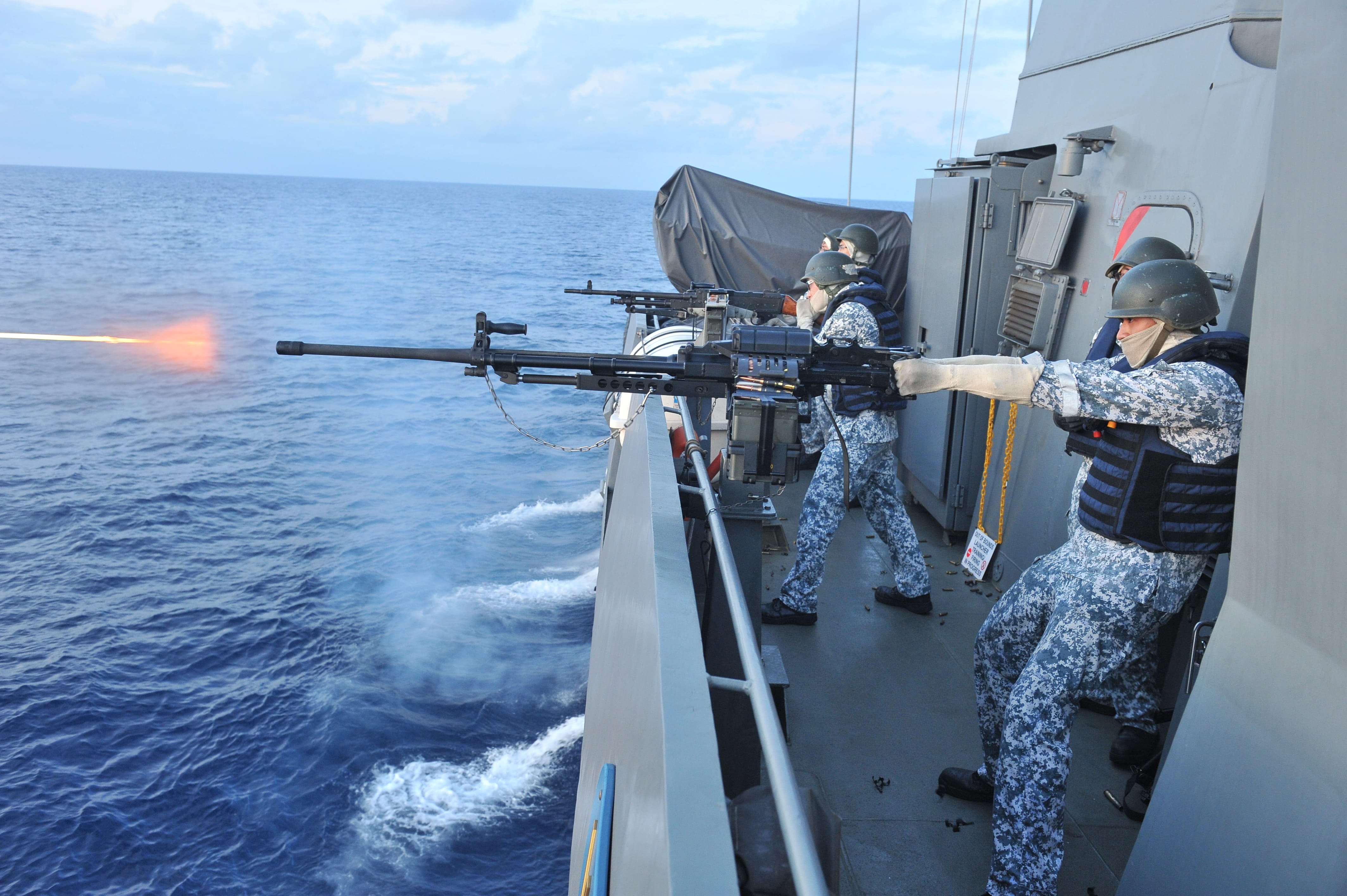 Navy personnel aiming a weapon from a ship's deck, with the open sea in the background.