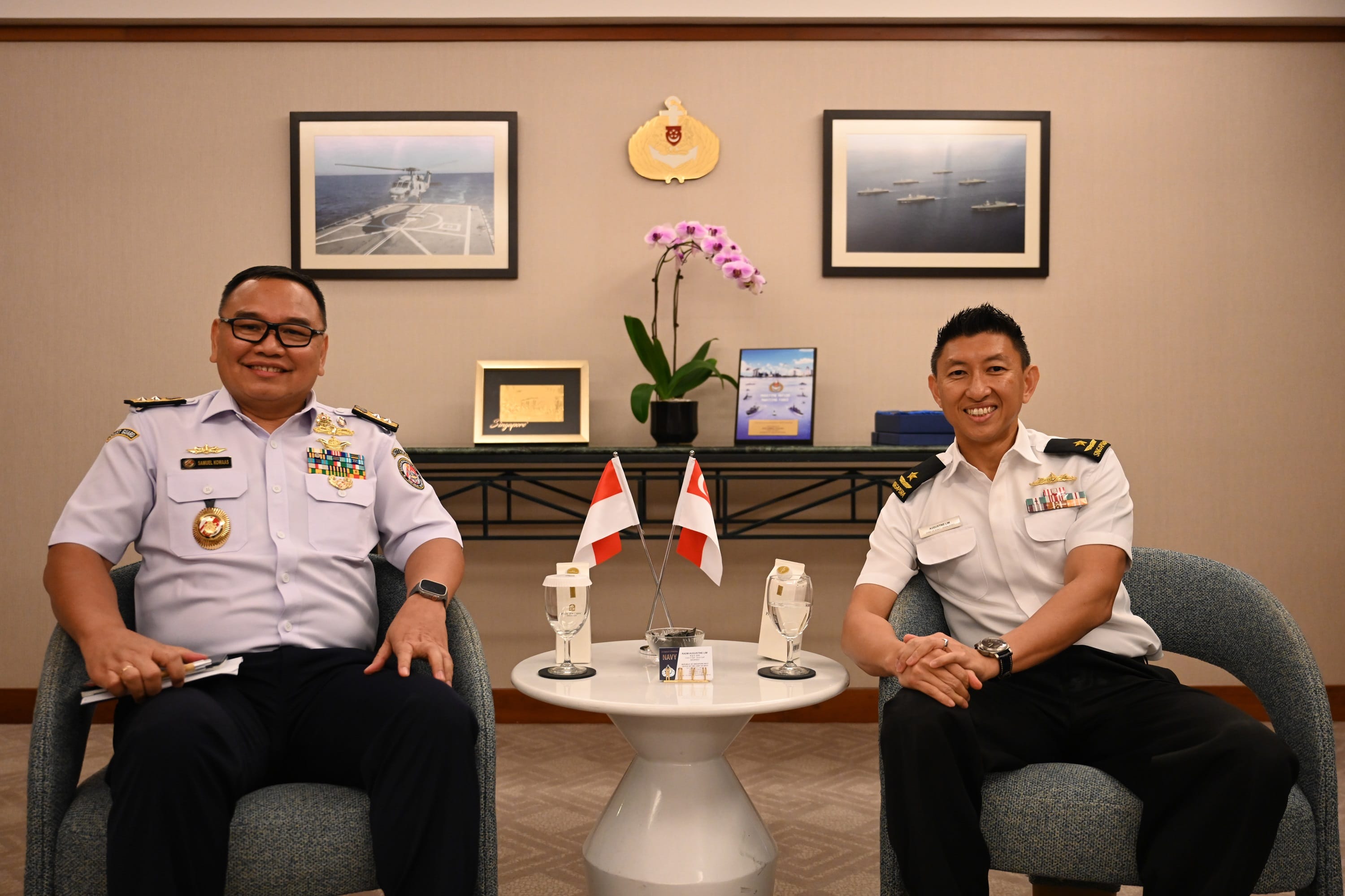 Two men in naval uniforms seated, with Singapore flags on table. Room has pictures of naval craft.
