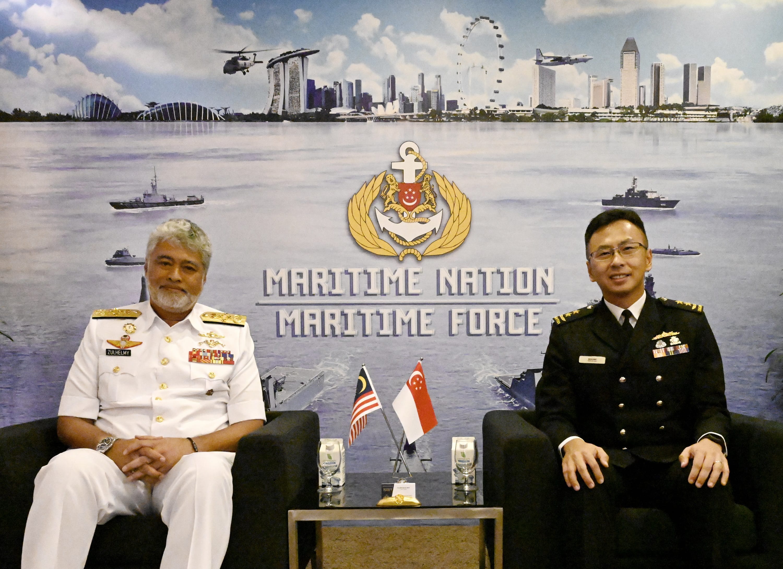 Two naval officers seated, with flags of Malaysia and Singapore in front, backdrop of Maritime Nation Maritime Force.