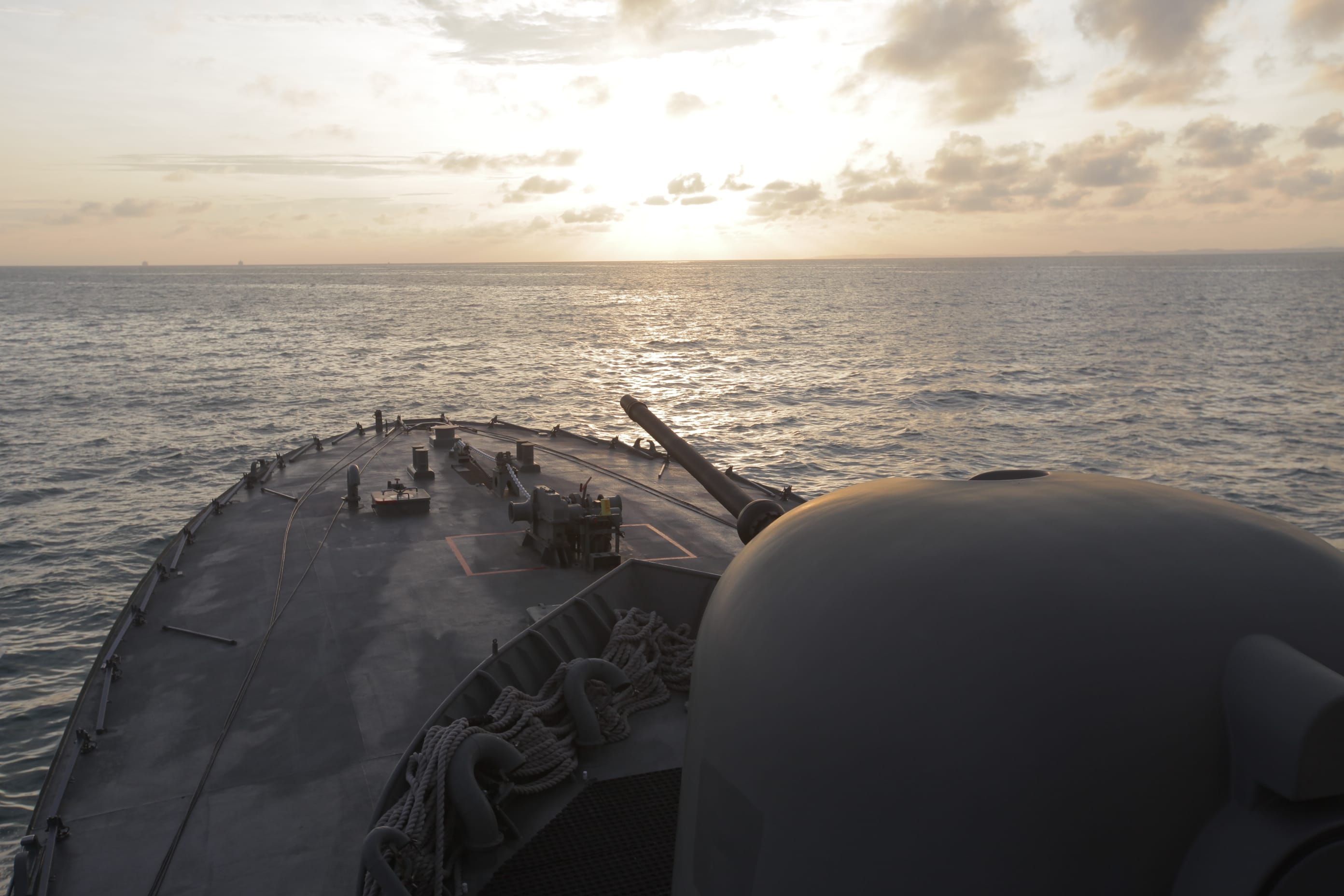 The bow of a naval vessel with a gun turret faces a sunset horizon over the sea.