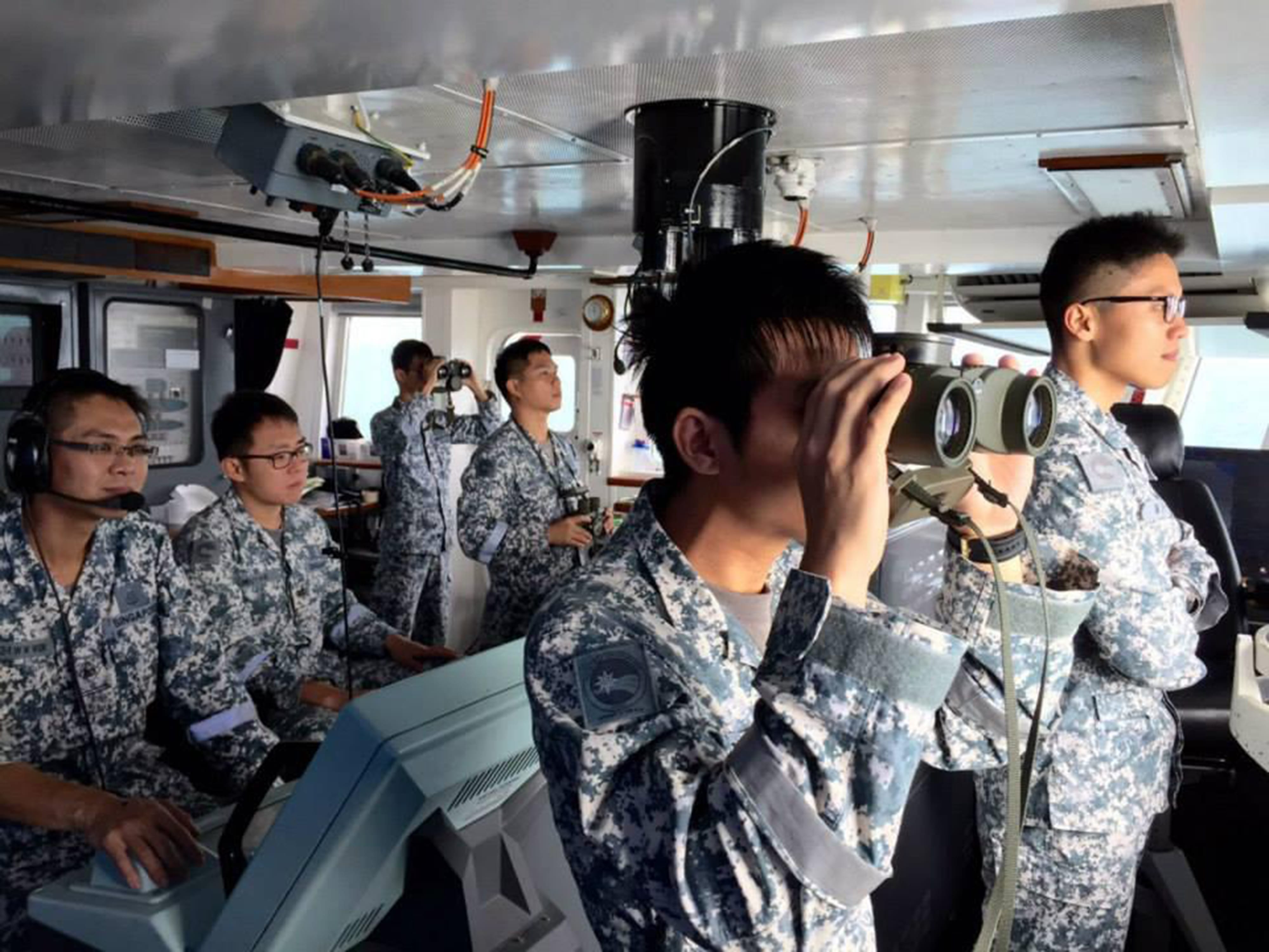 Sailors in camouflage uniforms monitor controls and use binoculars inside a ship’s bridge.