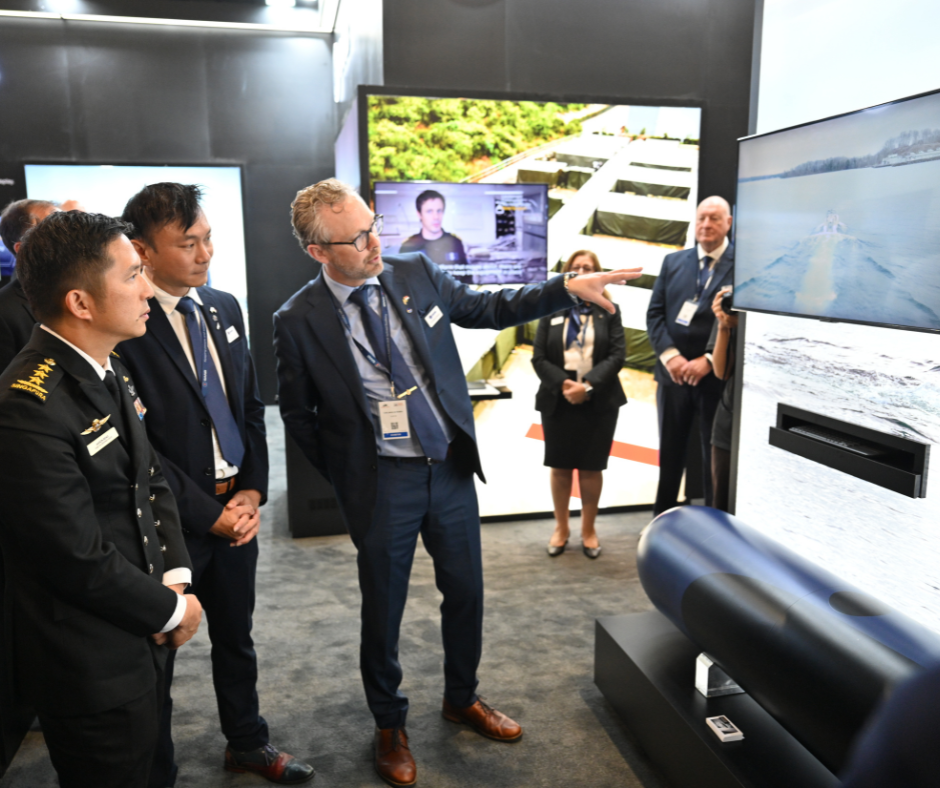 People at a trade show with a navy torpedo display and monitor screens.