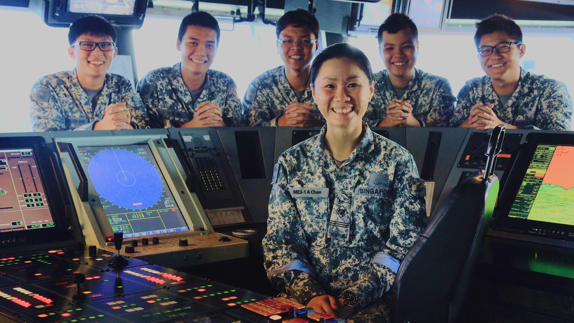 ME3 Angela Chan smiles in front of radar consoles on a naval ship's bridge with her team behind her.
