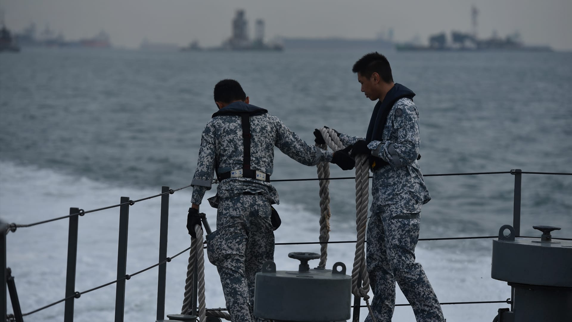 Naval personnel handling ropes on the deck of a ship with ocean background.
