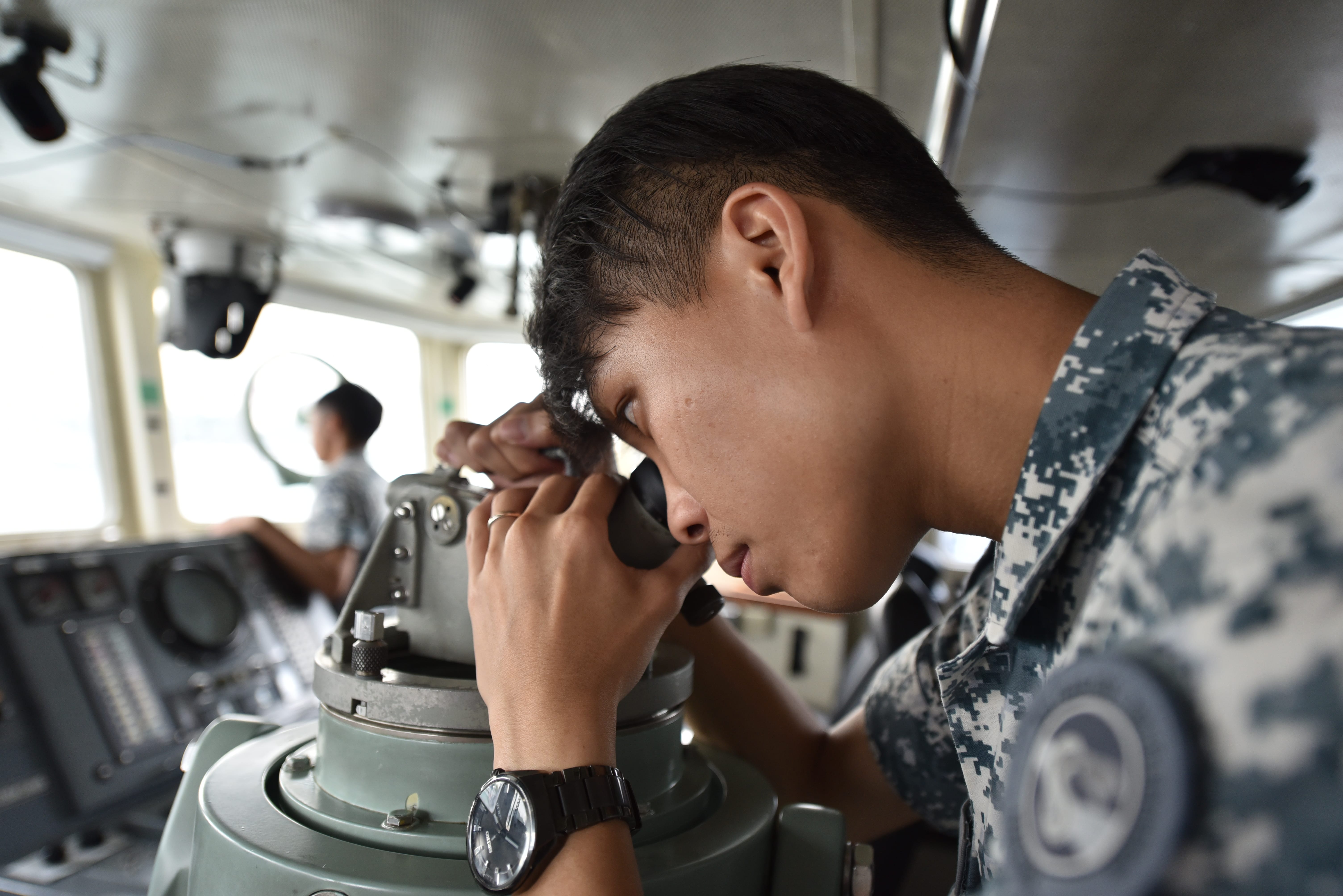 A naval crew member uses a periscope or optical instrument inside a ship's bridge.