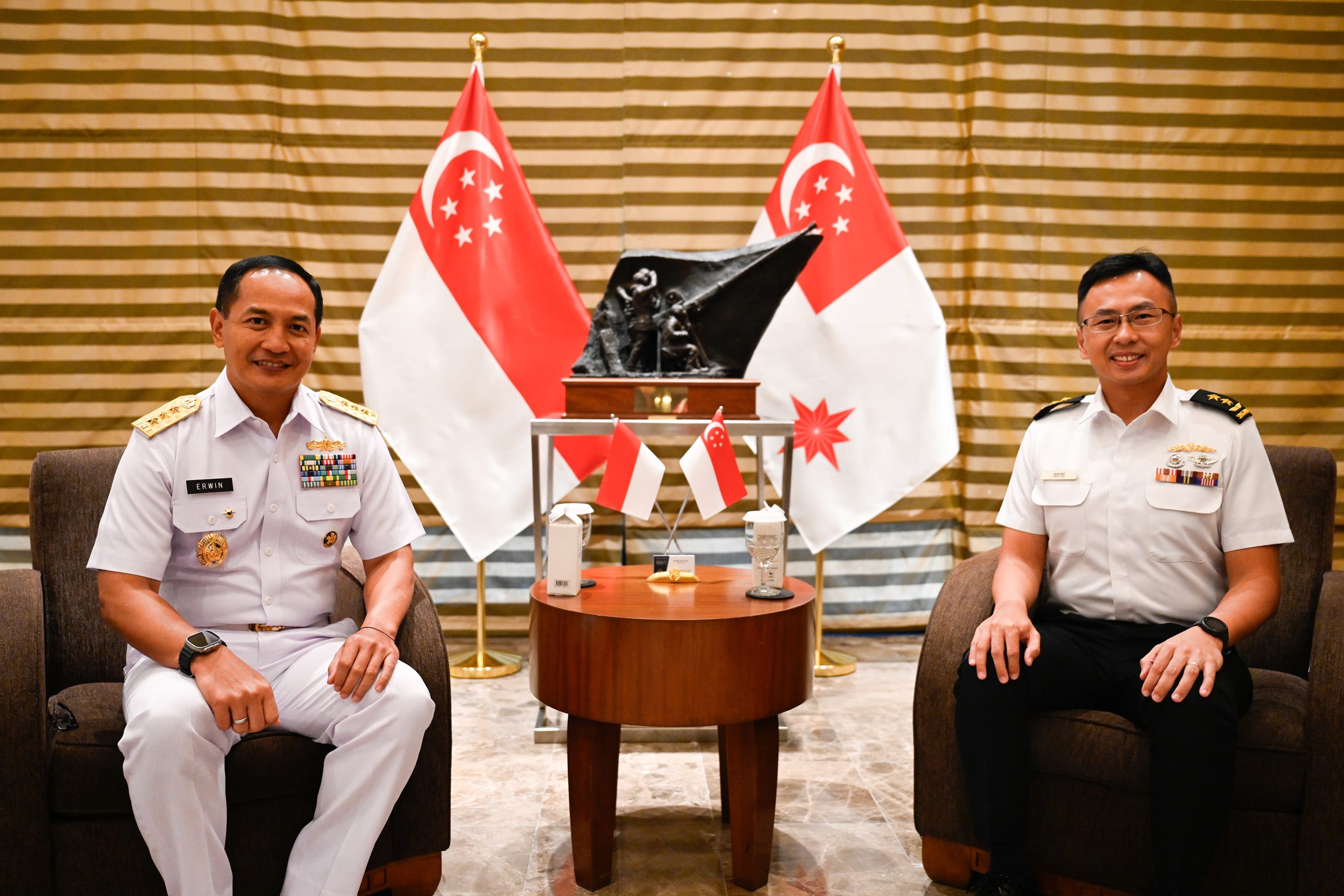 Two men in military uniforms seated, Singapore flags behind them.