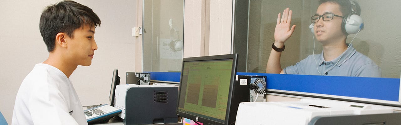 Man undergoing a hearing test in a soundproof booth, raising his hand while another person monitors results.