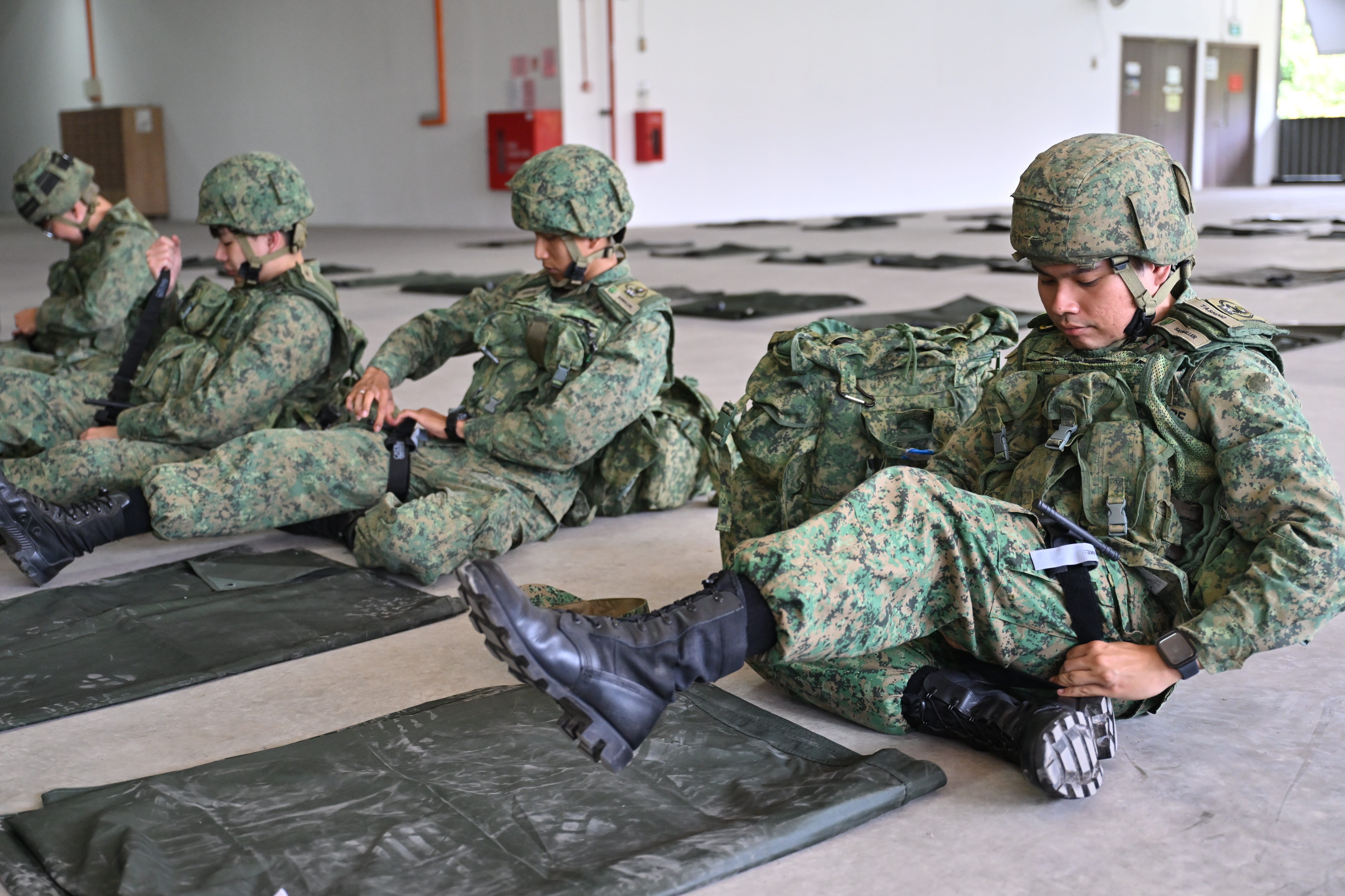 Soldiers in camouflage uniforms with helmets sitting on mats adjusting tourniquets.