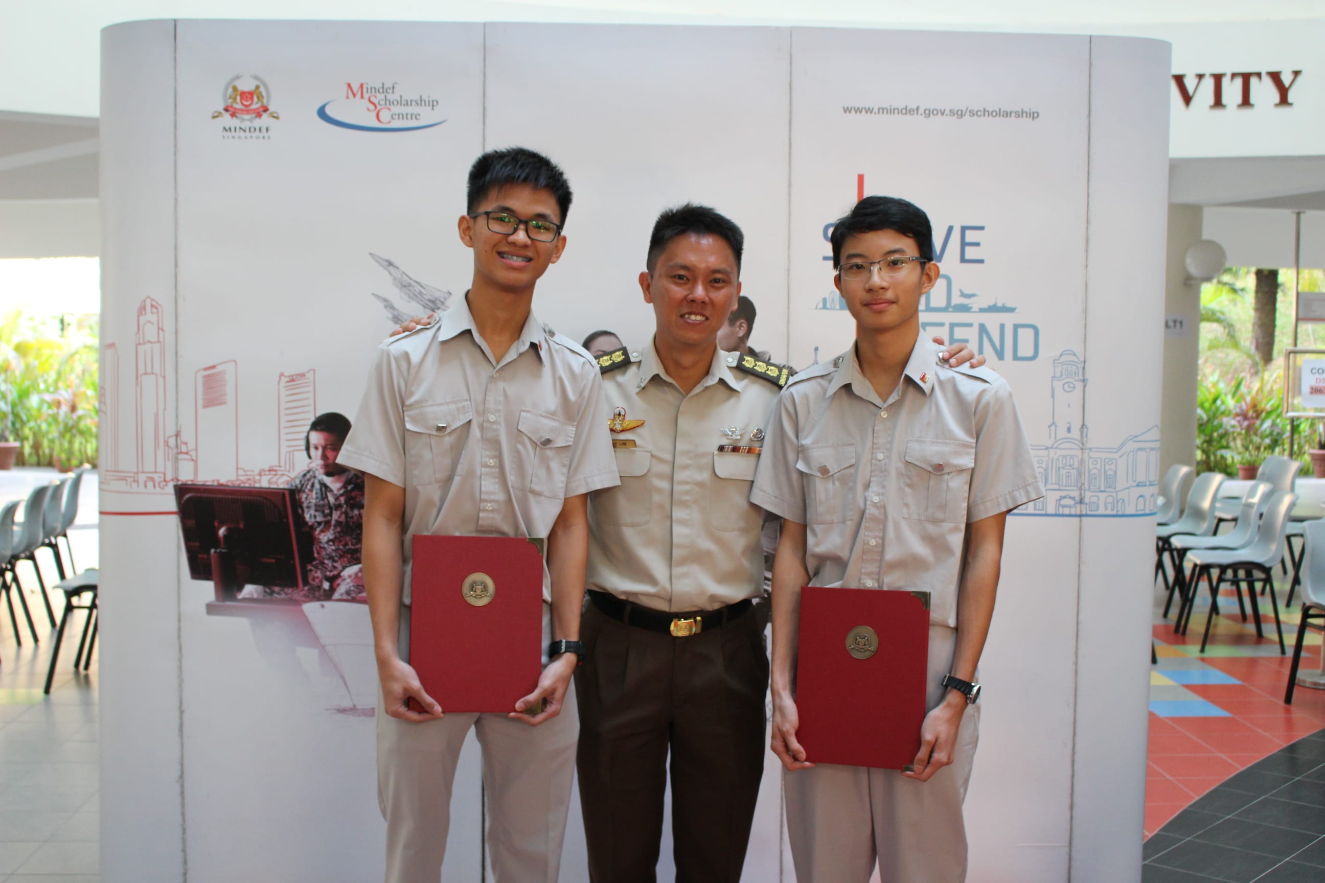 Two students holding red folders stand with one soldier in military uniform