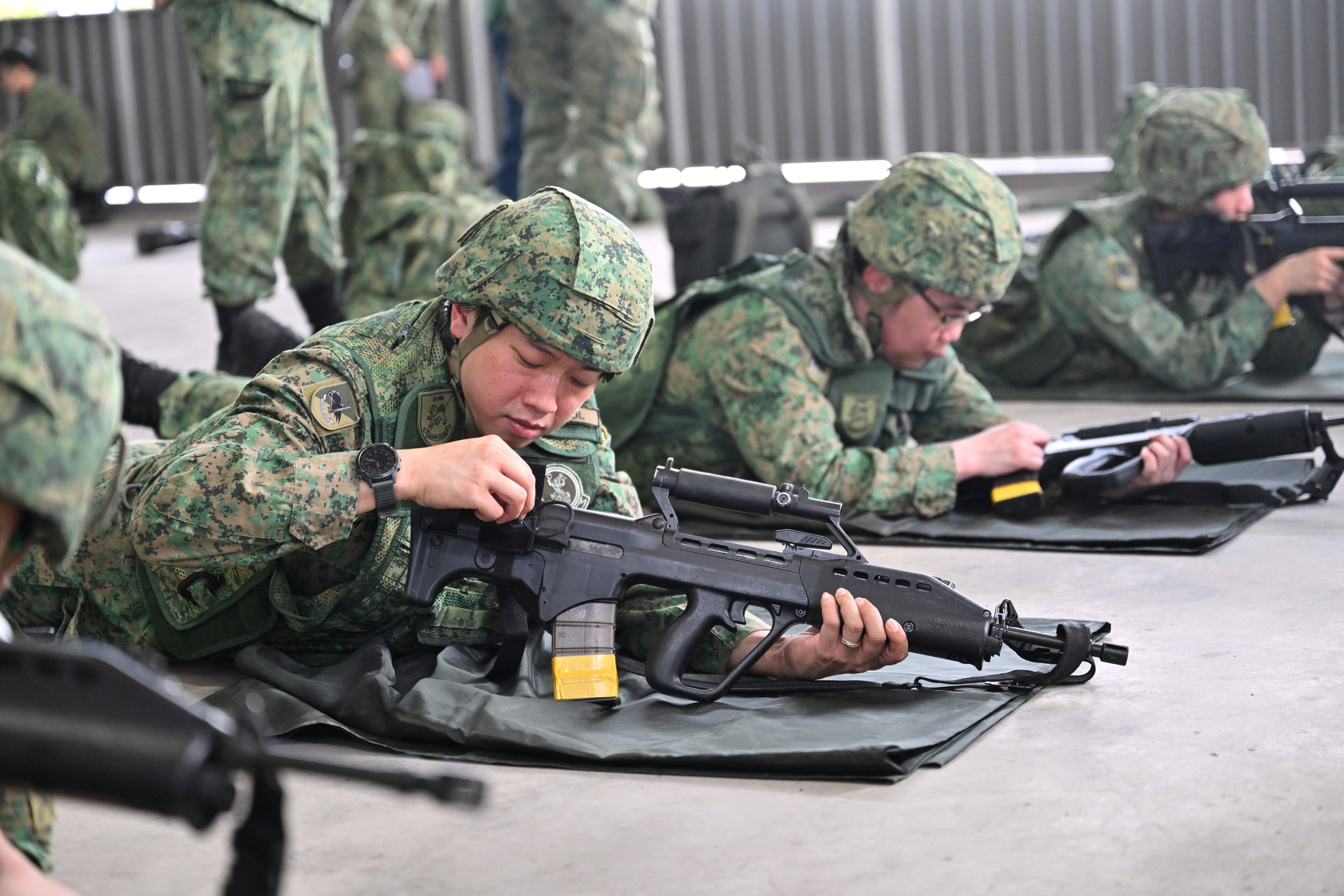 Soldiers in camouflage uniform lying down, handling SAR 21 assault rifles.