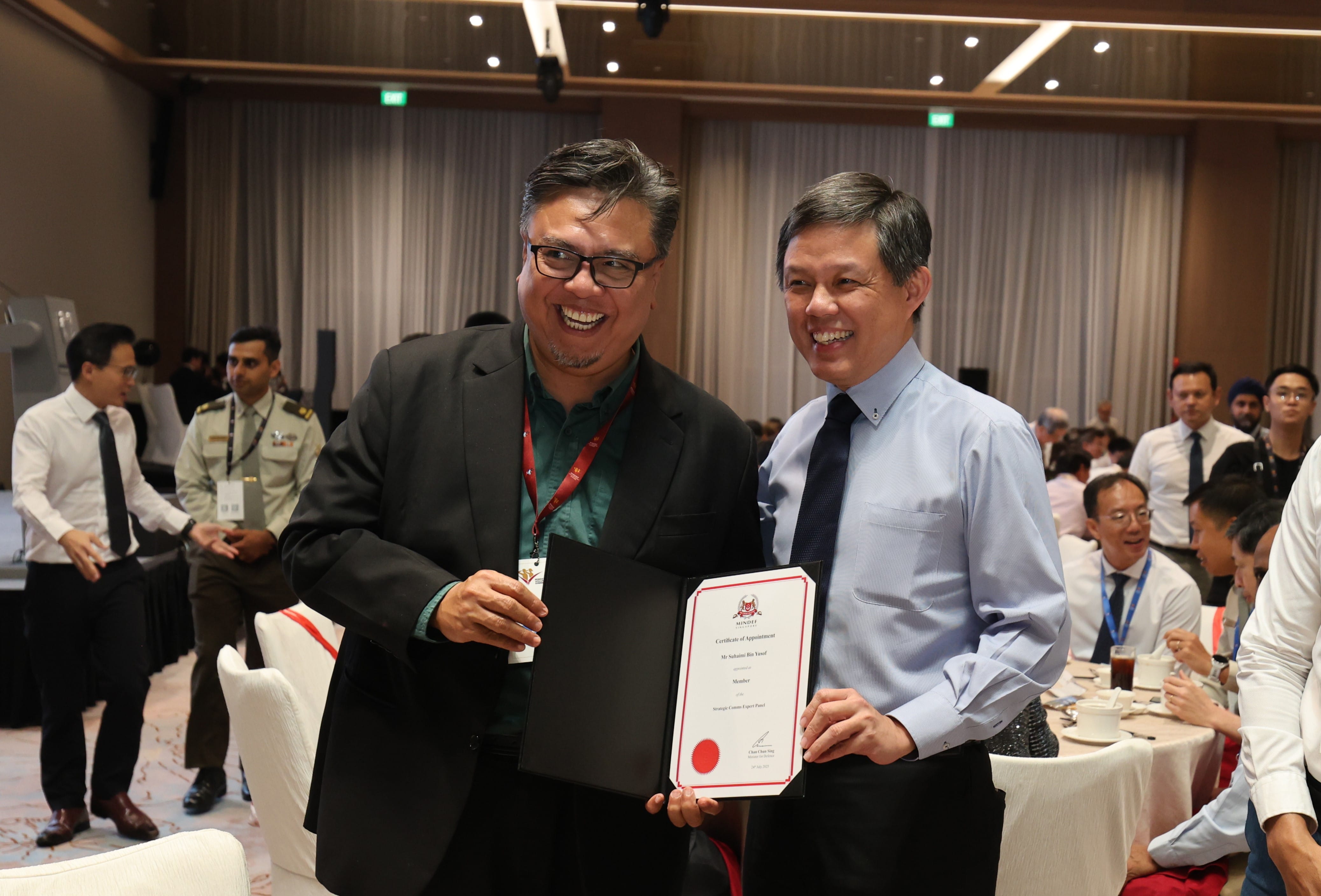 Two smiling men hold a MINDEF certificate of appointment; crowd in the background.