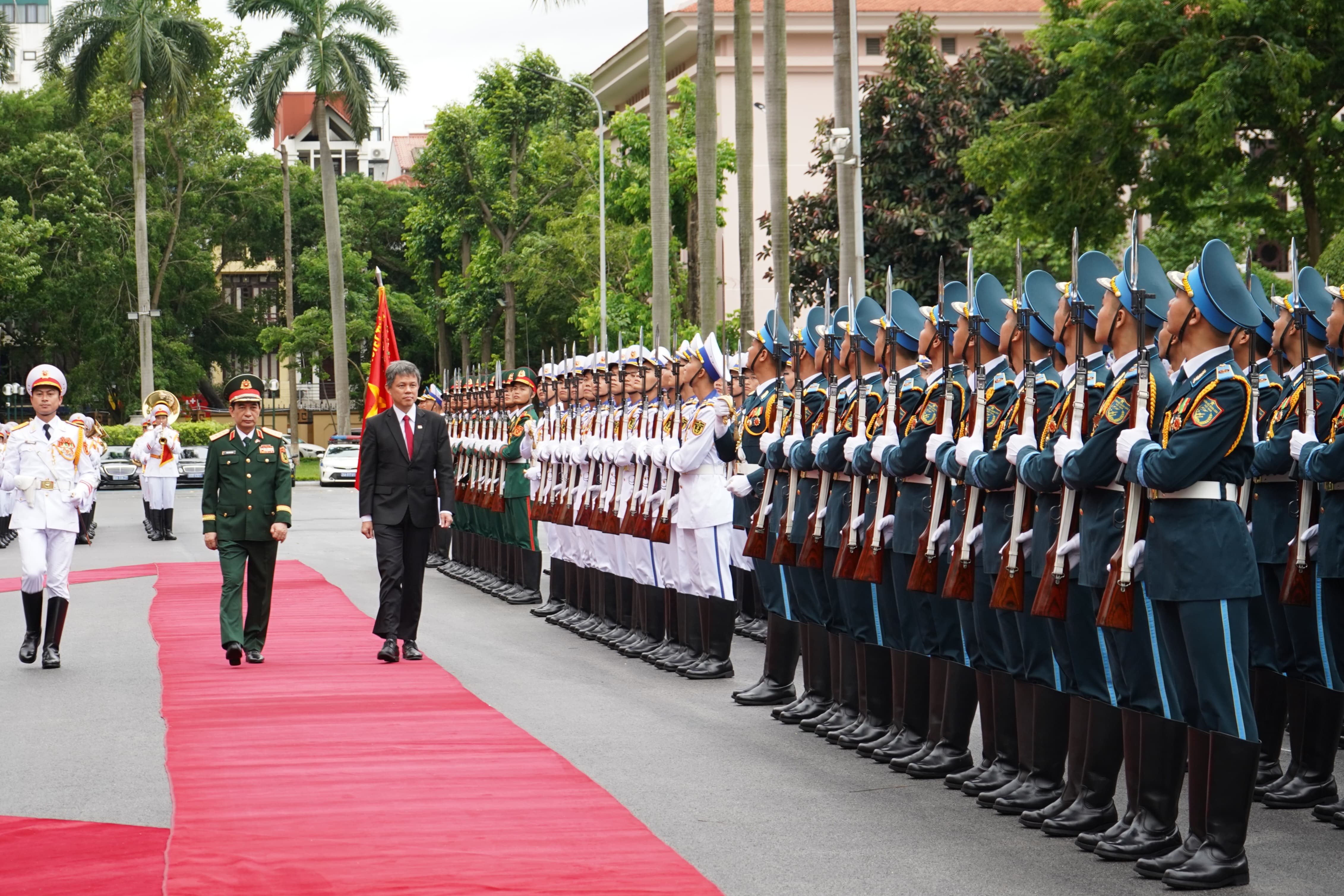 Men in suits and military uniforms walk on a red carpet past uniformed soldiers with rifles.