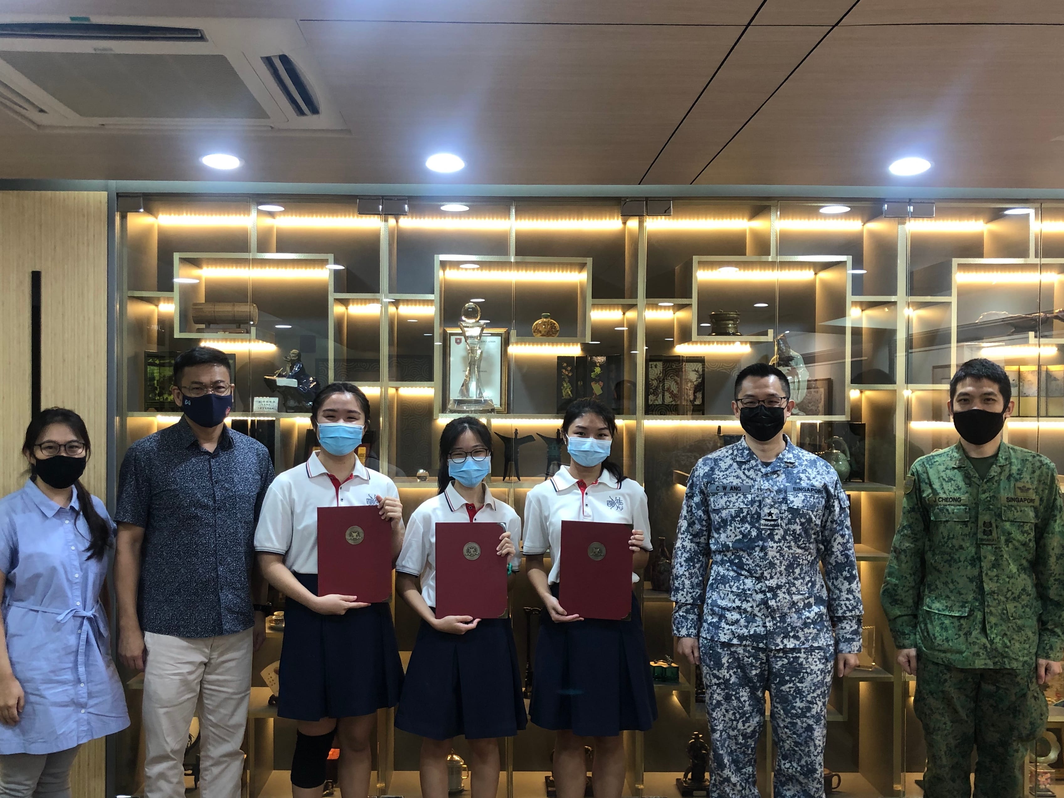 Three students holding red folders stand with four adults, two in military uniforms