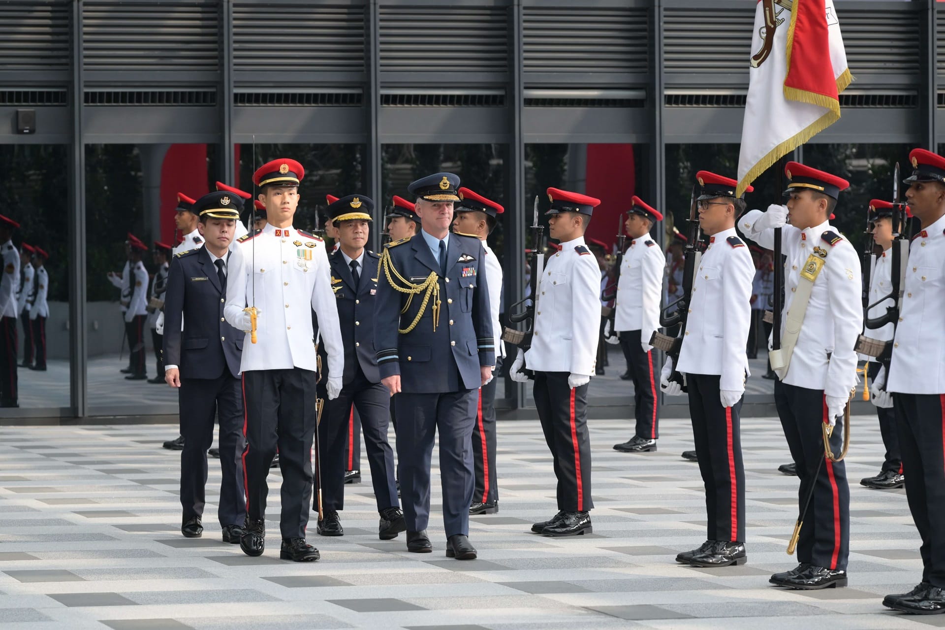 Military personnel in dress uniforms stand in formation outdoors. A flag is visible.