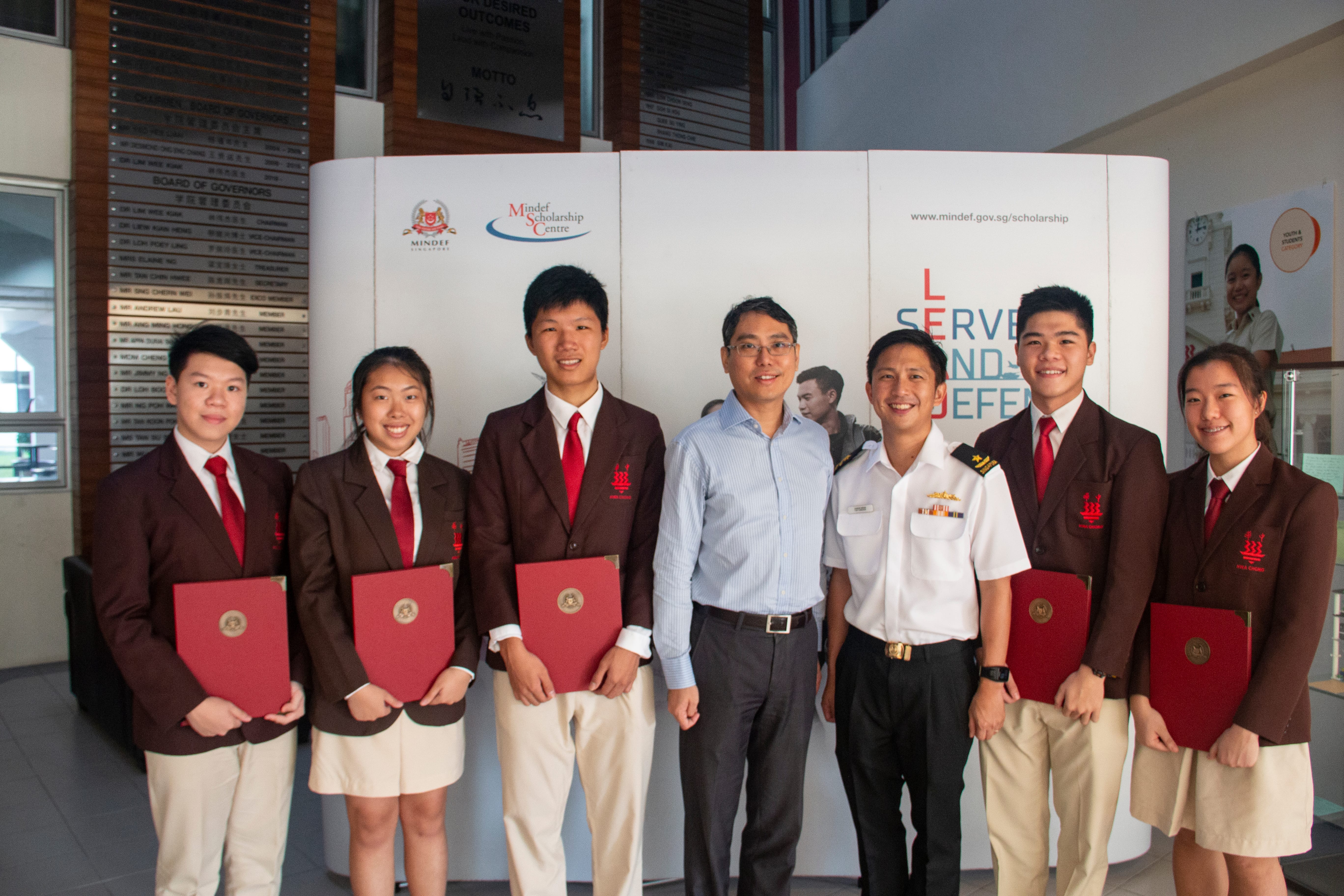 Five students holding red folders stand with two adults, one in military uniform