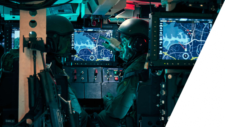 Inside of armored vehicle, two soldiers with helmets point at map screens.