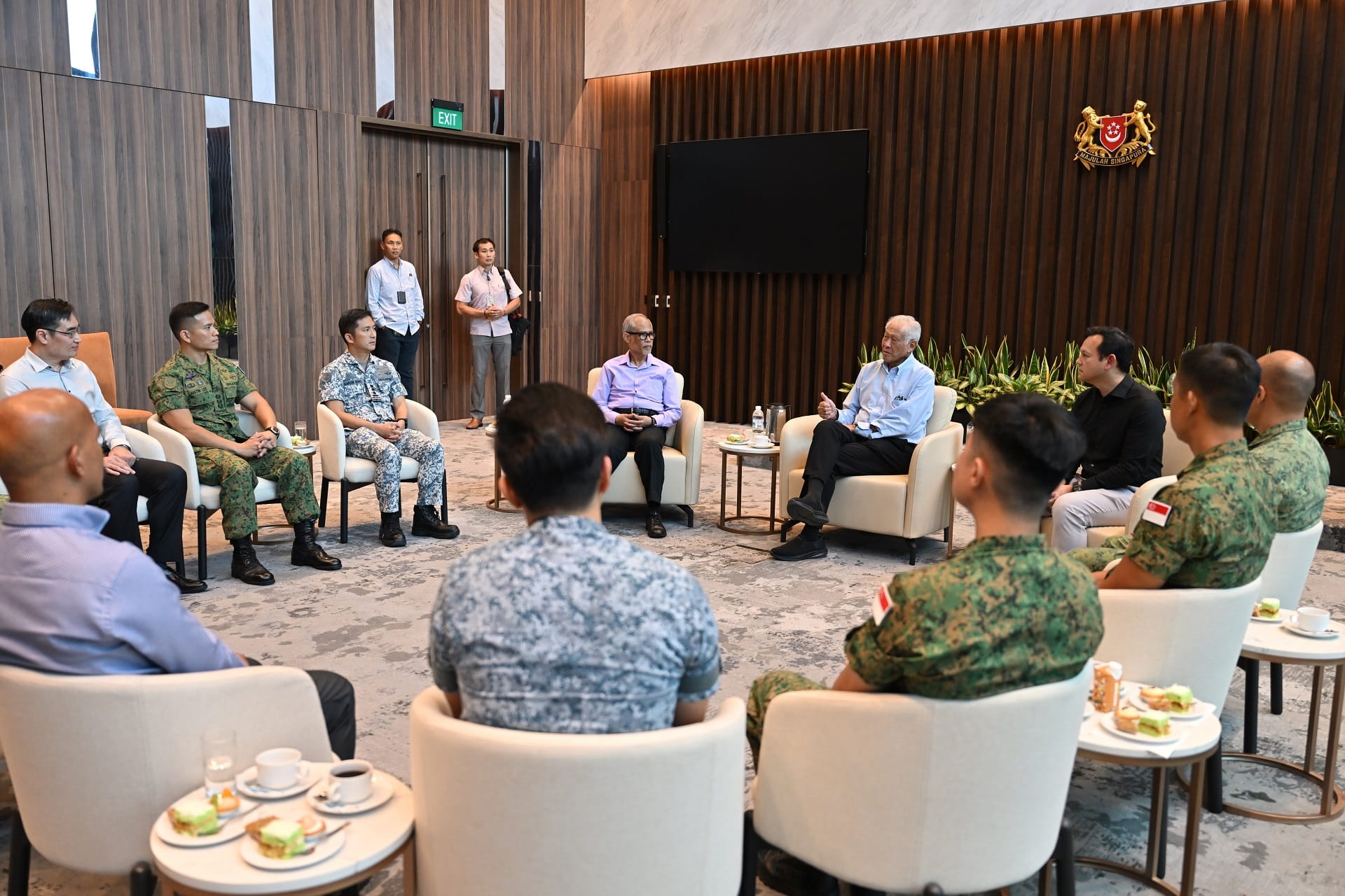 Meeting in room; Singapore coat of arms visible, plus soldiers in camouflage around two seated men.